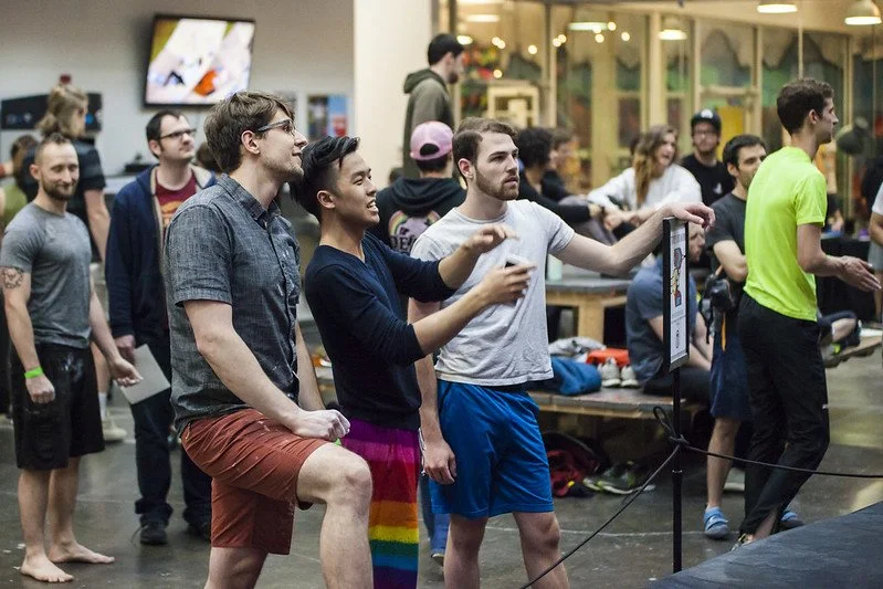 Group of people gathered indoors, watching and participating in a gaming event, with some playing video games on screens and others socializing.