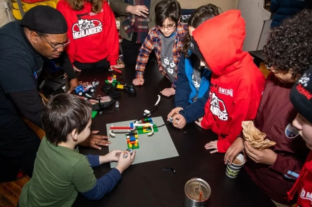 Group of children and an adult gathered around a table, working on a LEGO robotics project with colorful LEGO bricks, robots, and electronic components in a classroom or workshop setting.