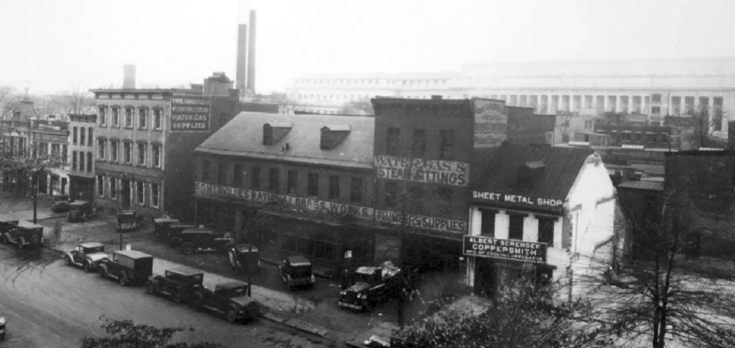 Black and white photo of a city street with old buildings, cars parked along the street, and advertisements for water gas supplies, sheet metal, and coppersmiths.