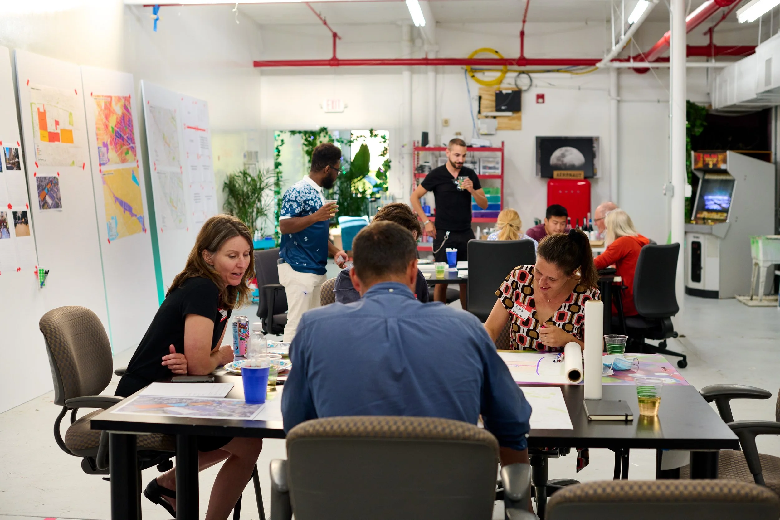 People gathered around tables in a bright, modern industrial-style room, engaging in a collaborative workshop or meeting, with posters on the walls and a large display showing the moon.