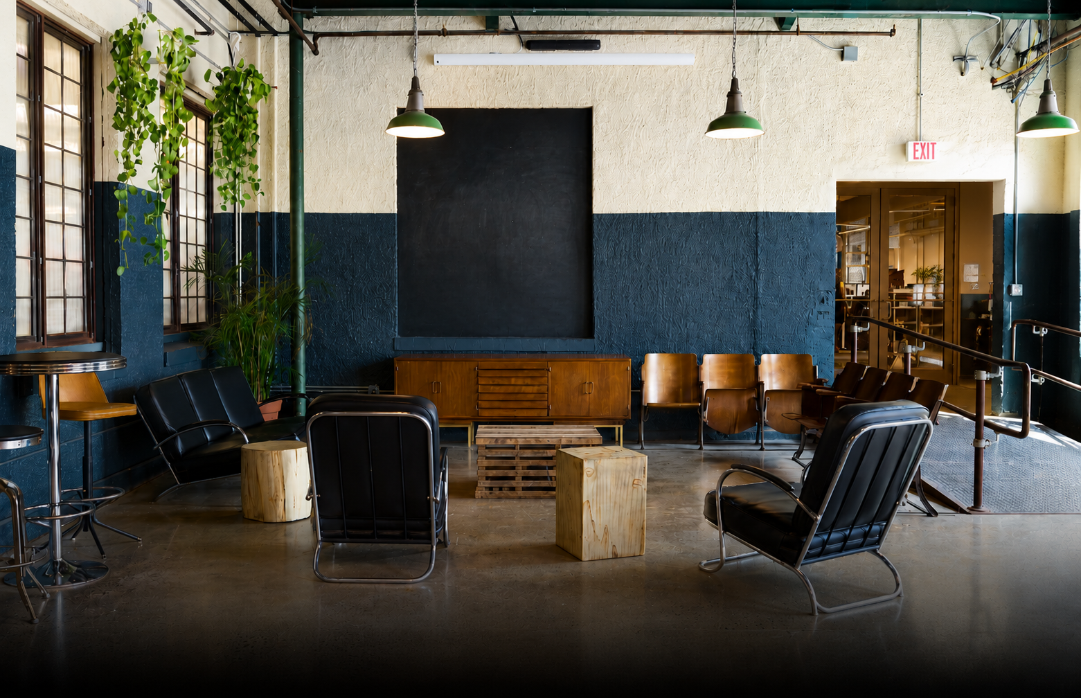 Indoor lounge area with black leather chairs and wooden seats arranged in a semicircle, a wooden cabinet, a black chalkboard, potted plants, and large windows with wooden frames, illuminated by hanging green lamps. The space has a textured beige and blue wall and a polished concrete floor.