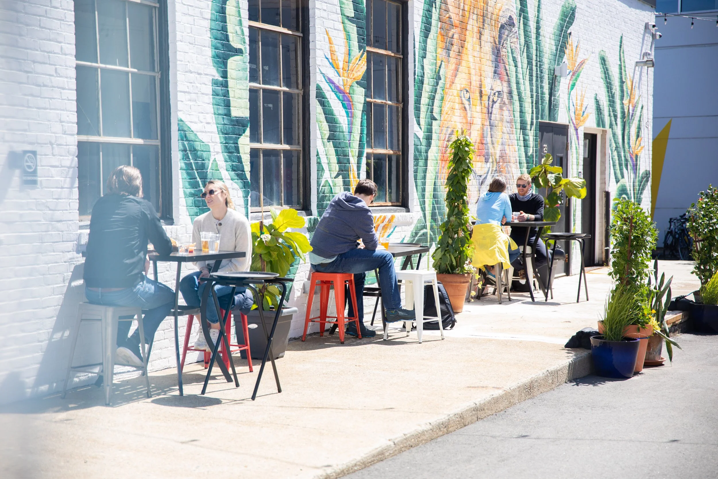 People sitting outside at tables with drinks in front of a white brick building with colorful mural and large windows