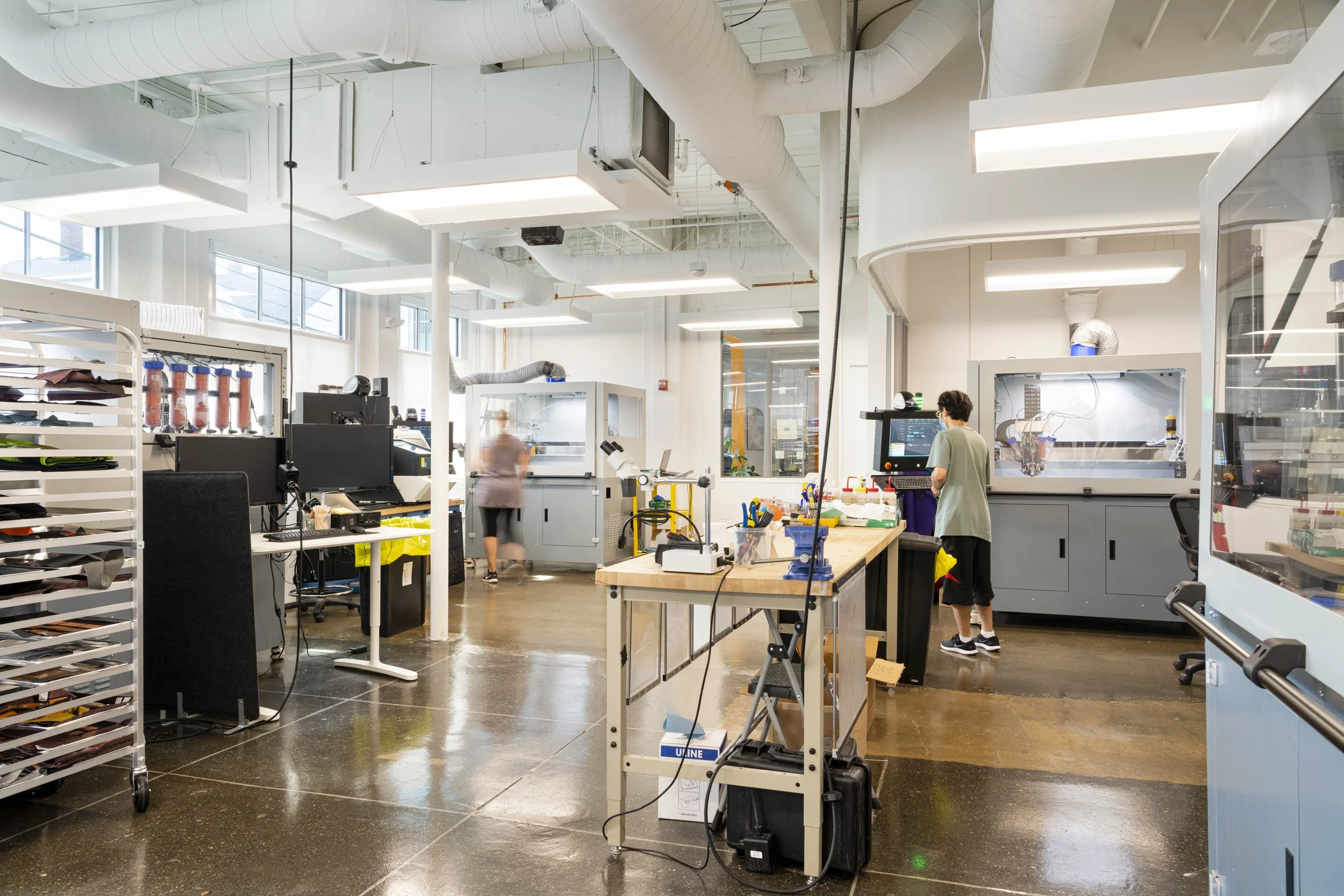 A modern industrial laboratory with large windows, white walls, and a high ceiling. There are workbenches, scientific equipment, and two blurred people working, including one person at a computer and another near a machine.