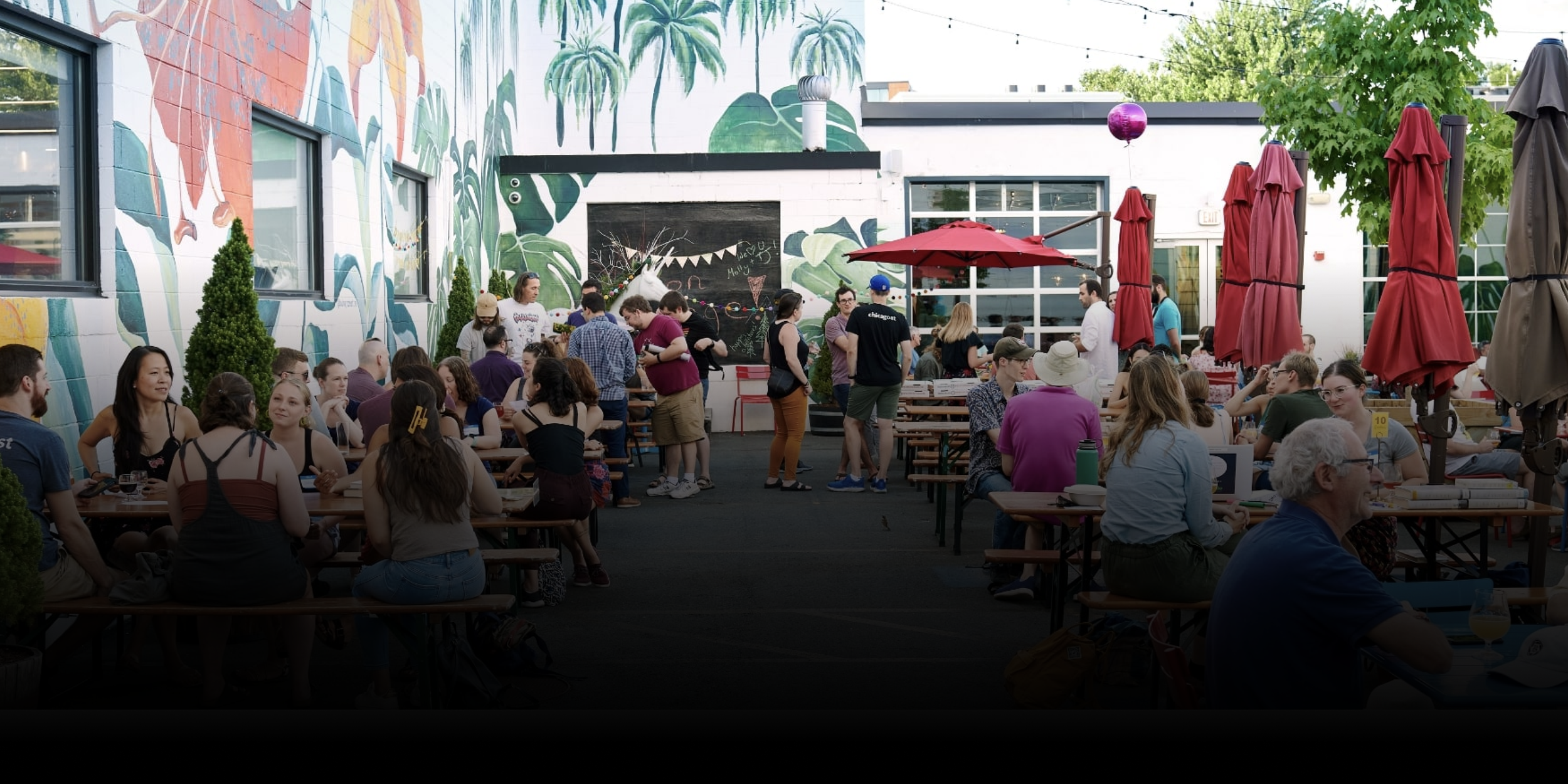 People sitting and standing at outdoor tables during a gathering at a patio with a colorful mural and greenery, with some individuals chatting and enjoying the event.