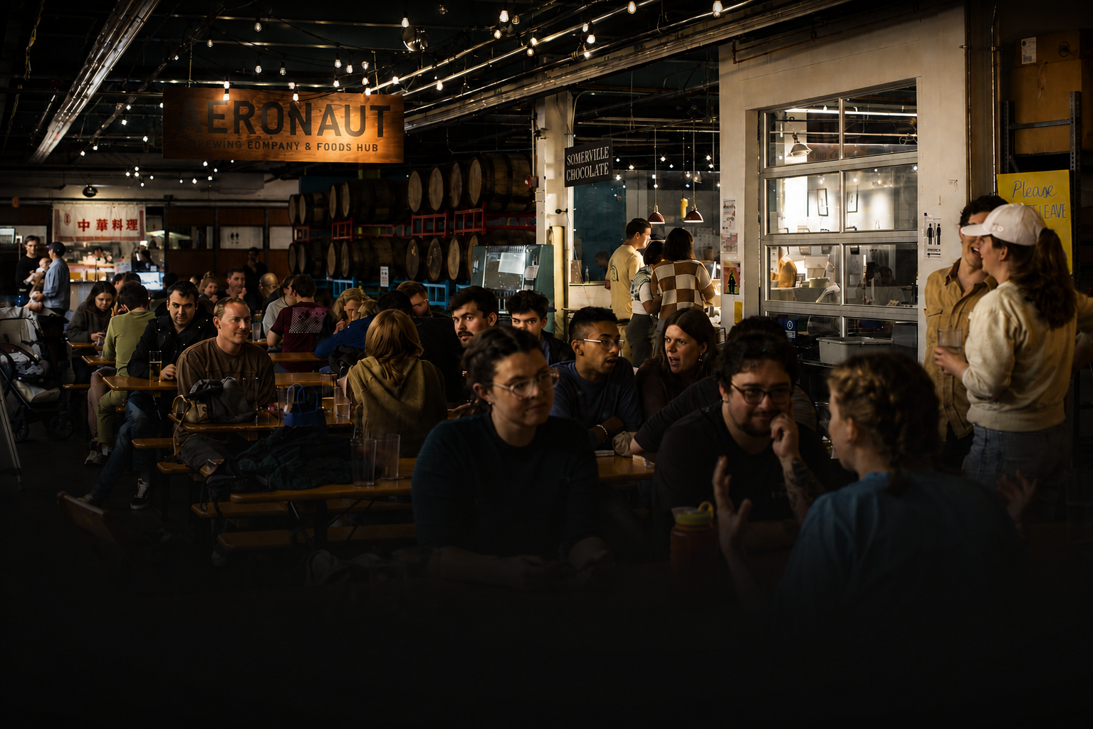 People dining and socializing inside a lively brewery with beer barrels and signages, including 'AERONAUT brewing company & foods hub' and 'Somerville chocolate', illuminated by string lights.