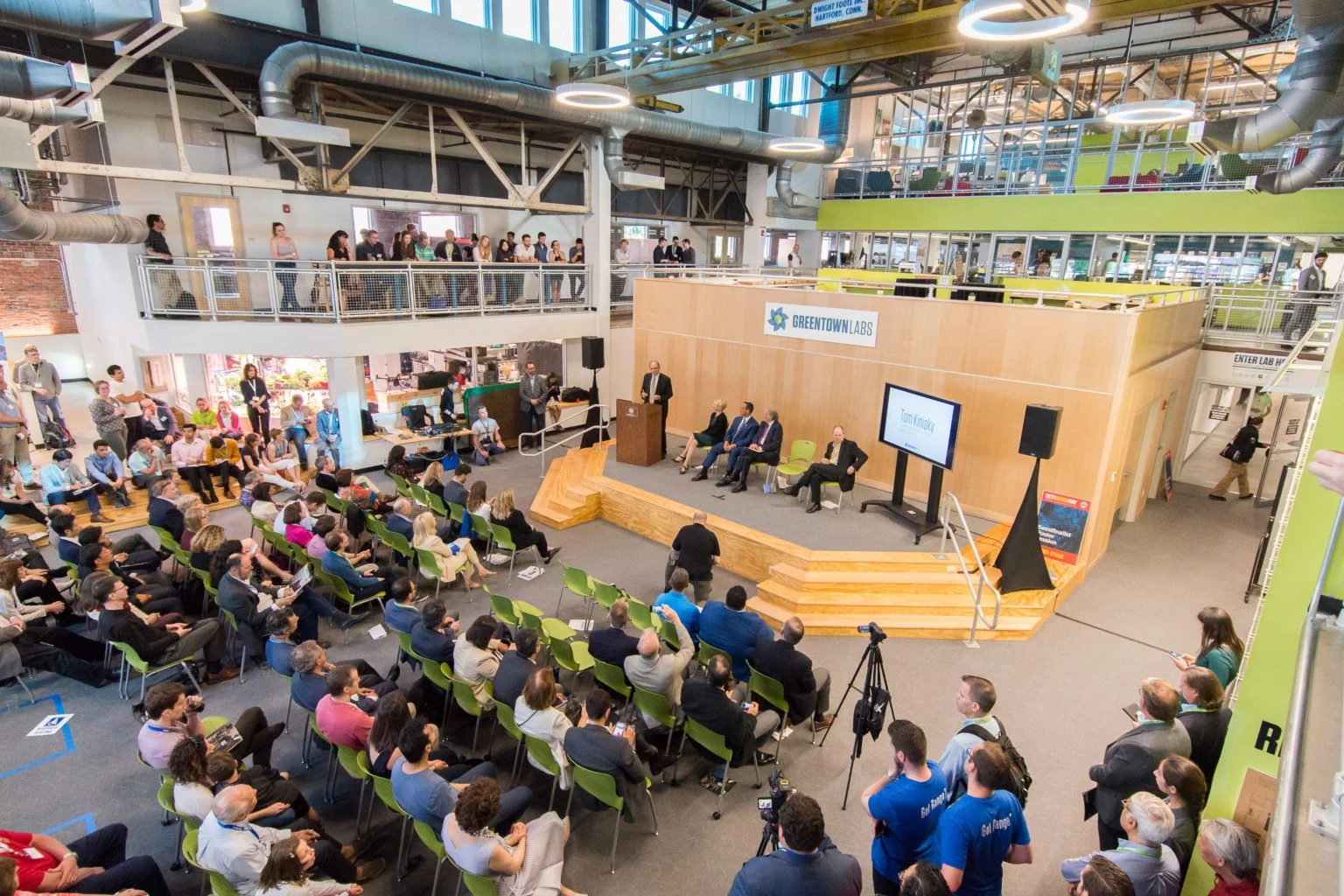 A large indoor conference or seminar with an audience watching a speaker at a podium on a stage. Behind the speaker, there are five seated panelists and a large screen displaying the name Tom Knesky. The room features multiple levels with people standing and watching. The venue has modern architecture with exposed ductwork and large windows.