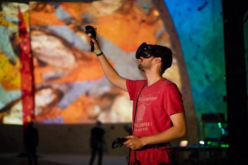 A man wearing a red t-shirt and a virtual reality headset holding a VR controller in his right hand, in front of a colorful abstract background.