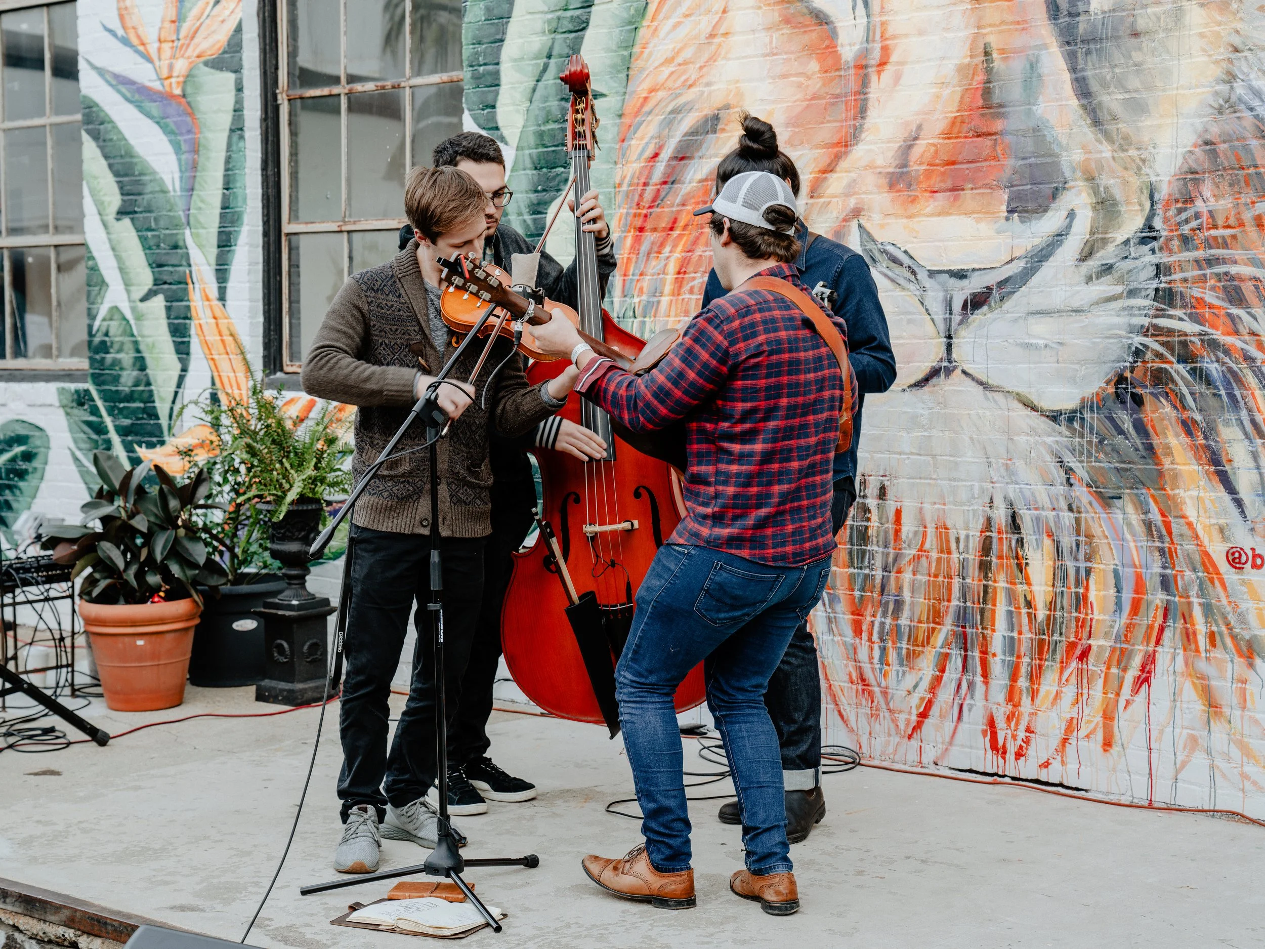 A group of five people playing musical instruments together outdoors in front of a colorful mural on a brick wall. One person is playing a violin, another is playing a double bass, and a third is playing a guitar. They appear to be performing or practicing.