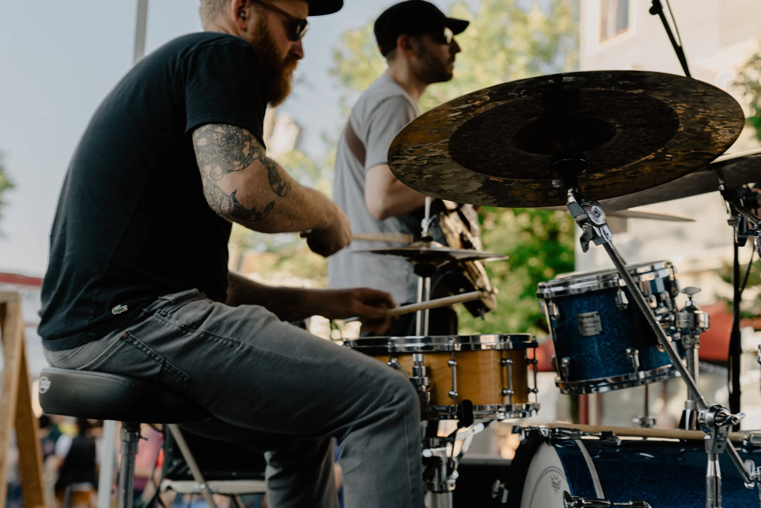 Two men playing drums outdoors during the daytime. One man is wearing sunglasses, a black t-shirt, and has a tattooed arm. The other man is wearing a gray t-shirt and a black cap. Both are focused on playing the drums, with a background of trees and a building.