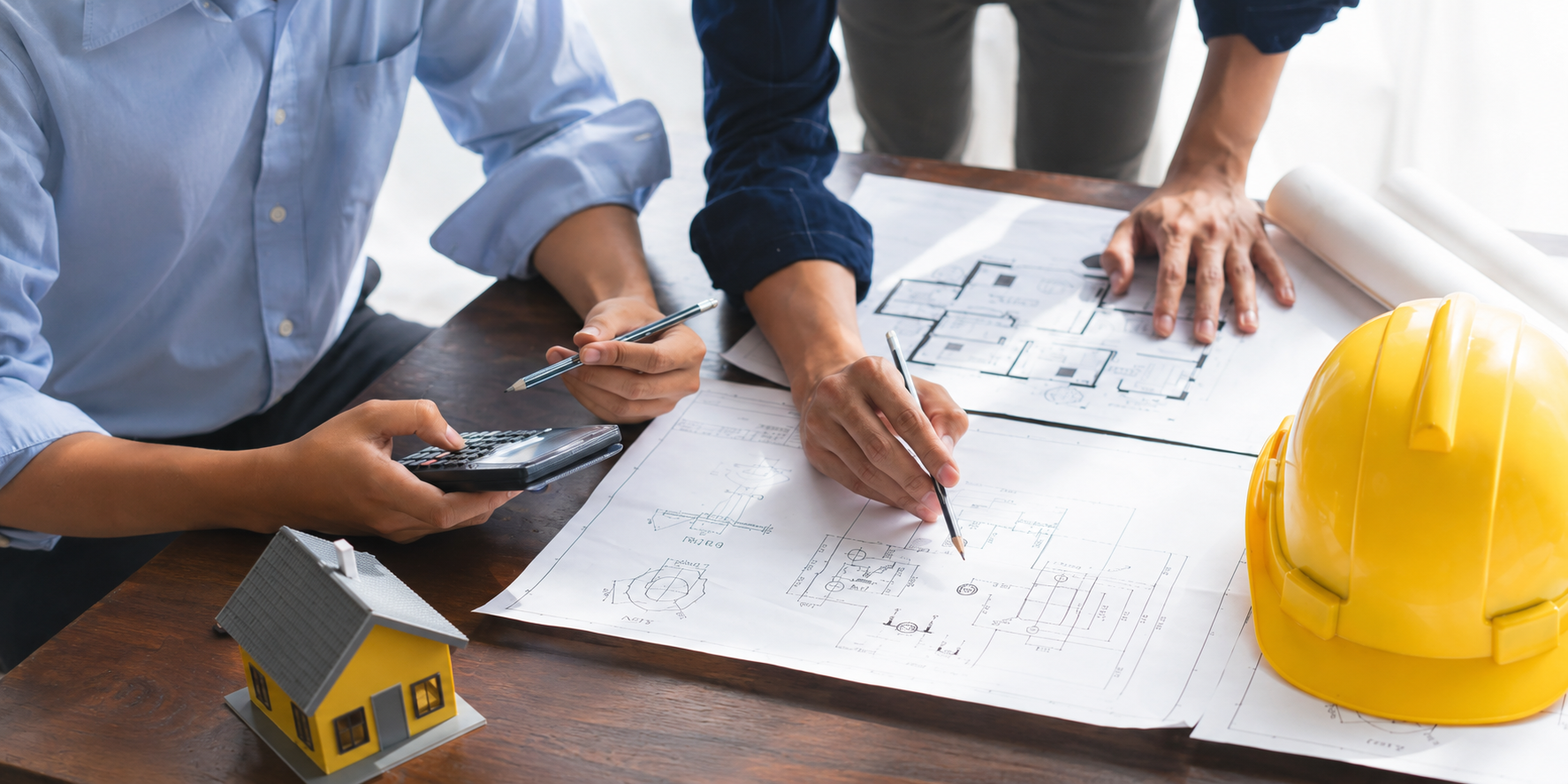 Architects or engineers reviewing blueprints on a wooden table with a yellow construction helmet and a small model house.