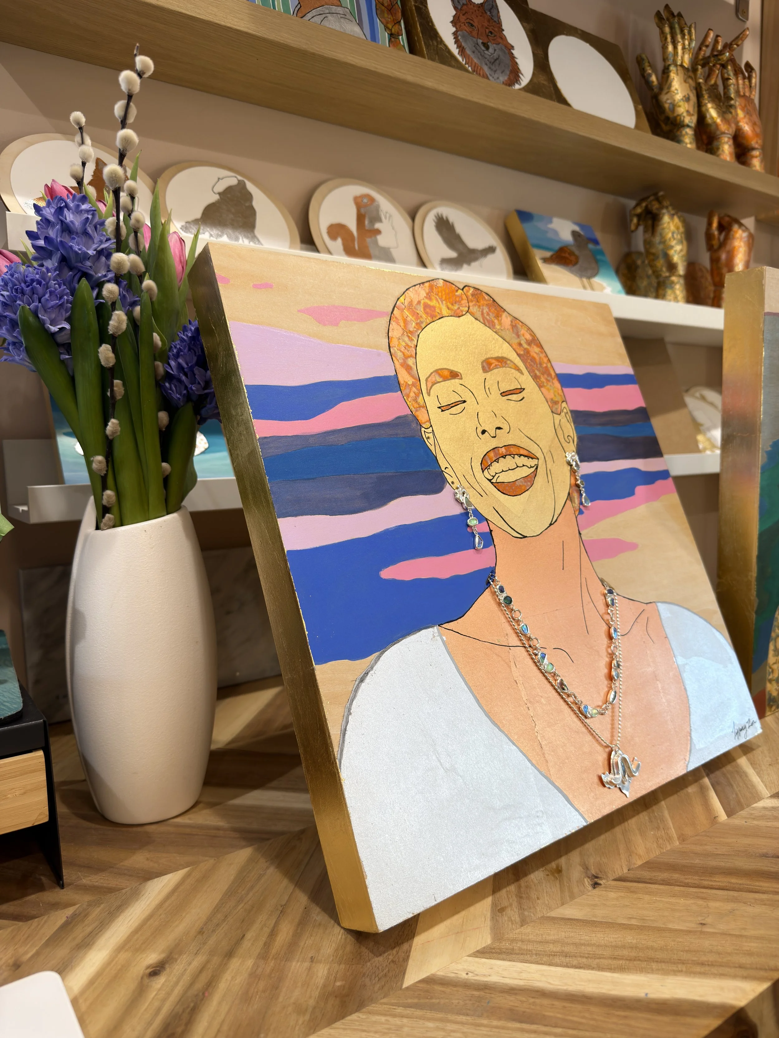 A colorful portrait painting of a woman with short hair, earrings, and necklace, set against a sunset or sunrise background with pink, purple, and blue sky. The painting is on a wooden easel and is placed next to a white vase with purple flowers.