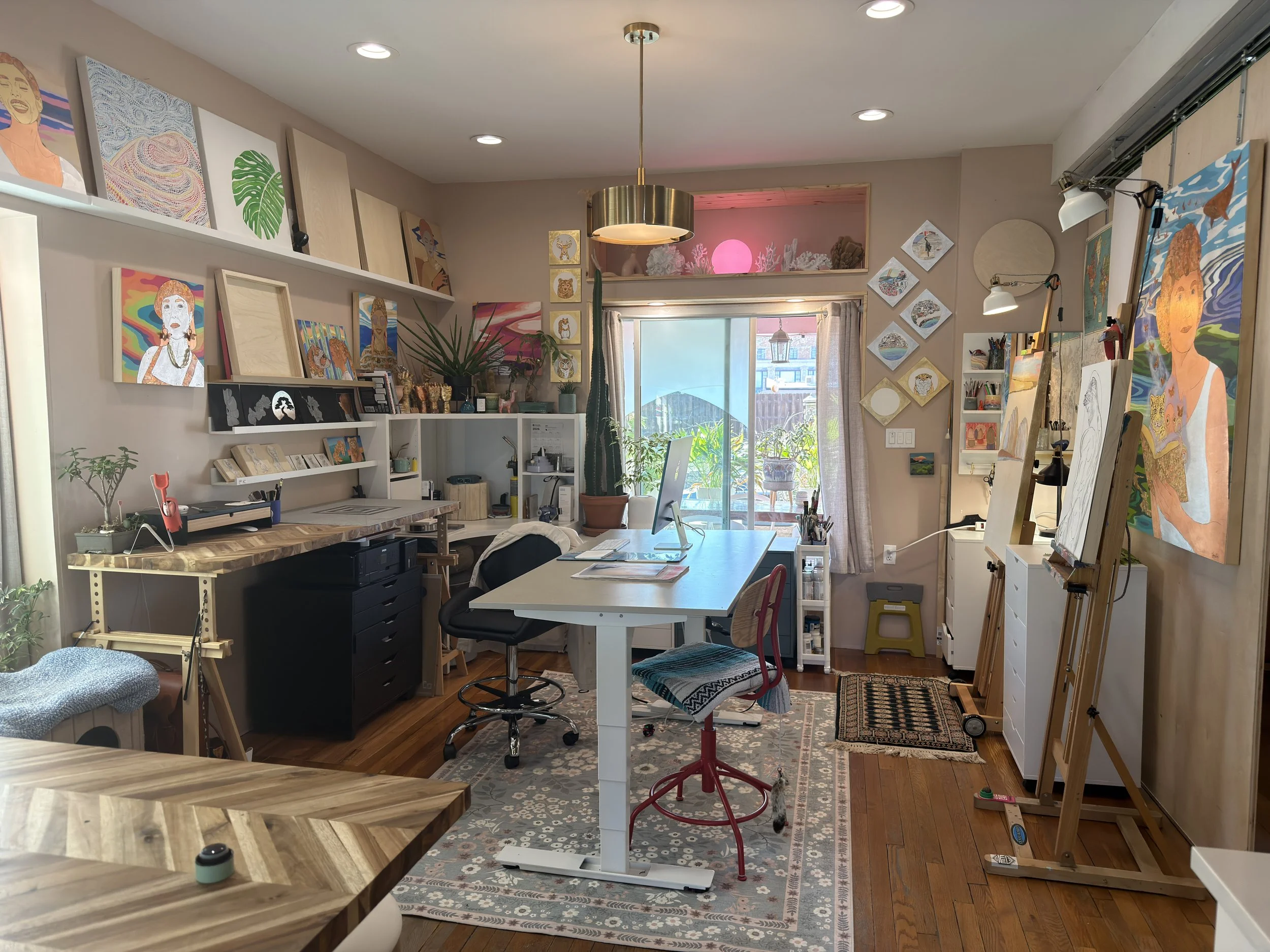 A cozy art studio with paintings on the walls and shelves, a large window with curtains, a desk with a computer, chairs, and various art supplies and tools.