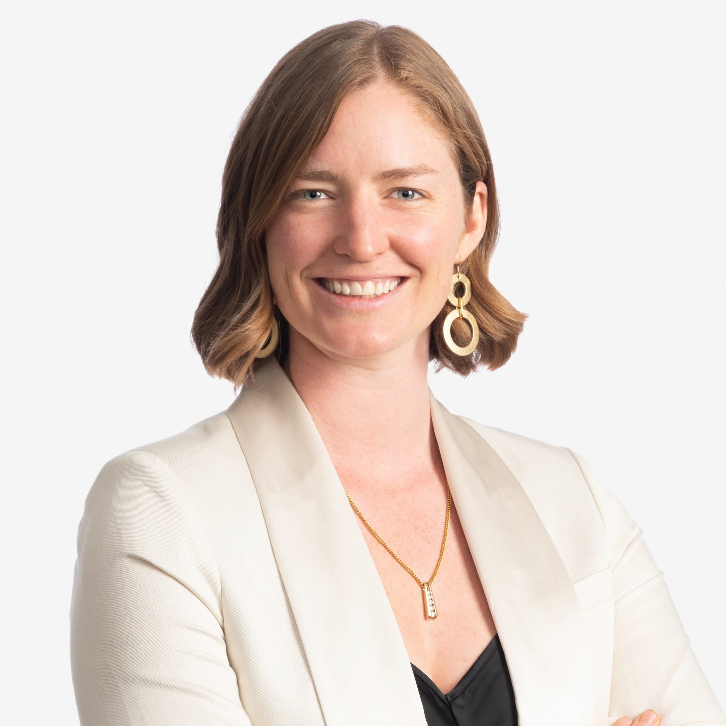 A woman with shoulder-length brown hair smiling, wearing gold earrings, a gold necklace, a black top, and a white blazer, against a plain white background.