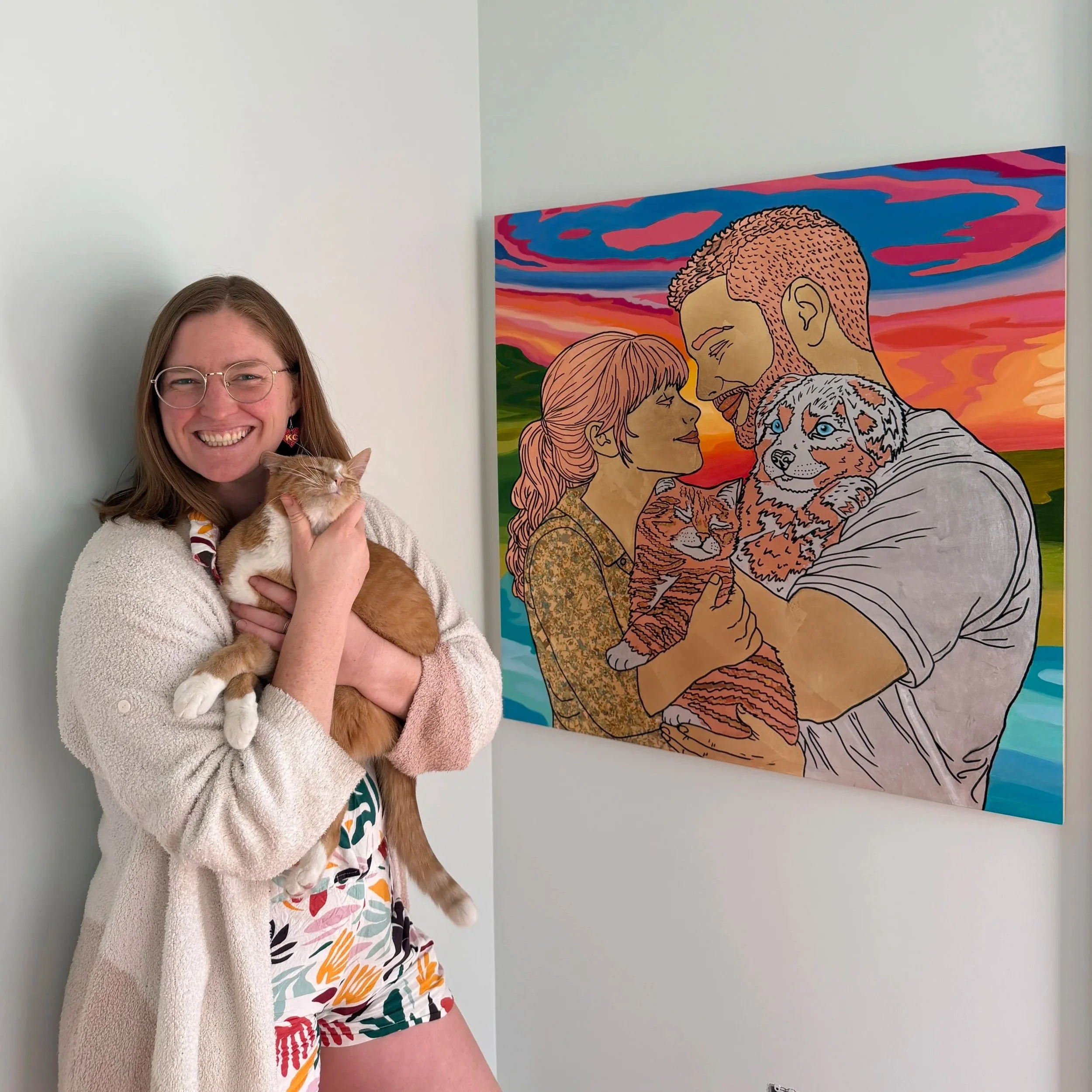 A woman with glasses and long hair smiling while holding a ginger and white cat. She is wearing a patterned dress and a light-colored cardigan. Behind her is a colorful painting of a couple holding two cats, with a sunset background.