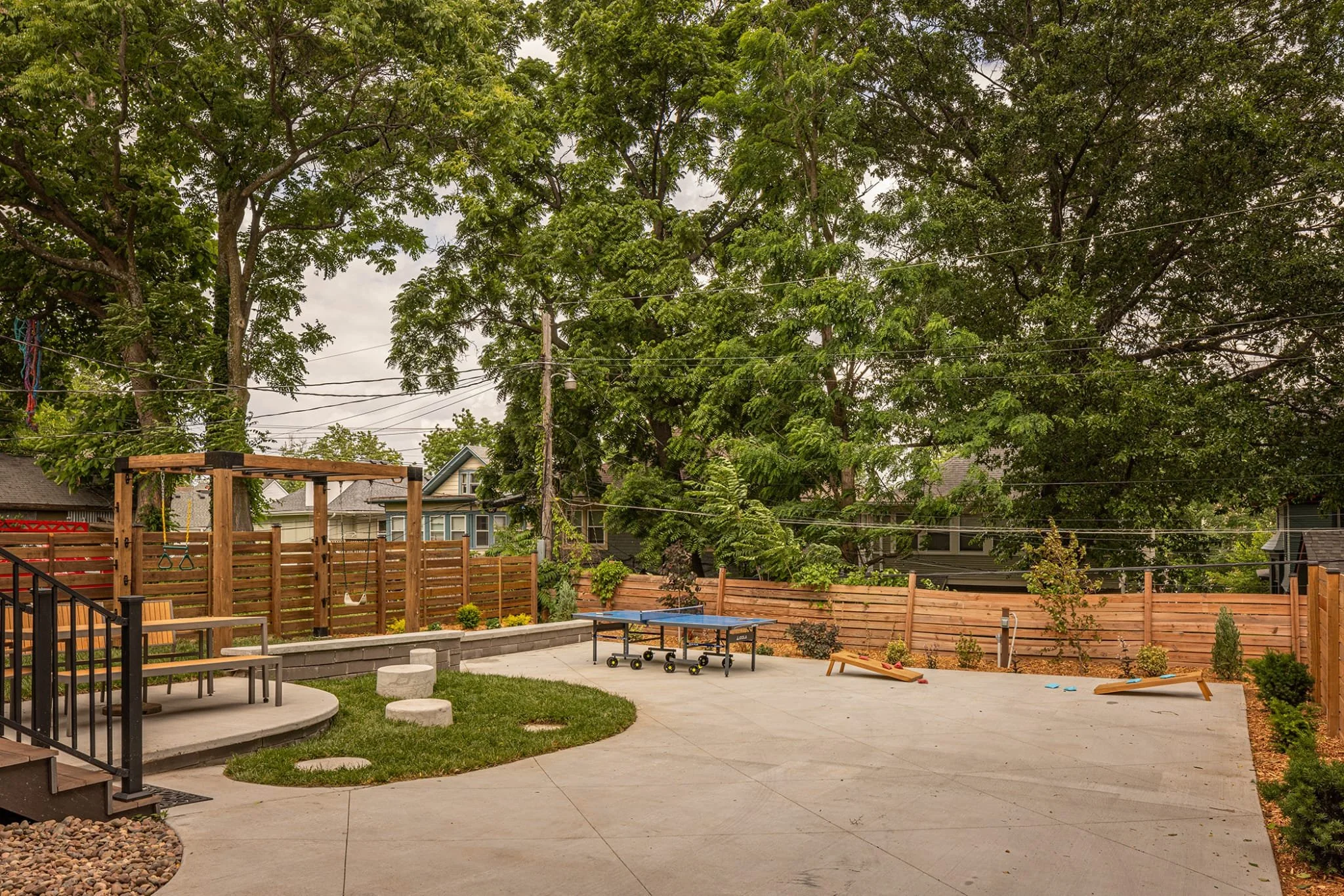 View of a backyard patio with a concrete surface and a landscaped flower bed along the fence, containing small plants and mulch, with a wooden fence and trees in the background.