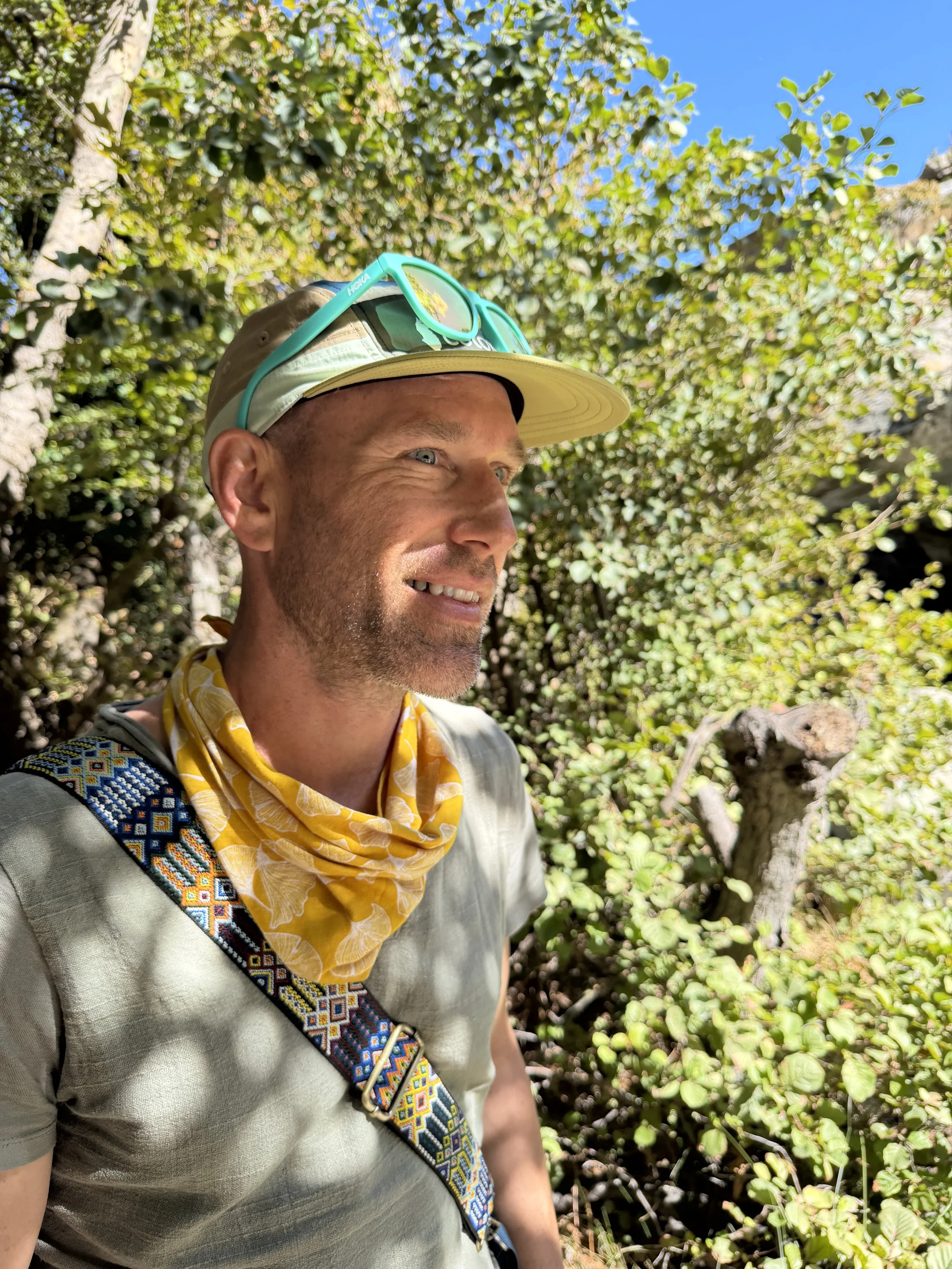 A smiling man with a beard and blue eyes outdoors in sunny weather, wearing a beige cap, green sunglasses, a yellow patterned bandana around his neck, and a gray T-shirt with a colorful strap across his chest, surrounded by green foliage.