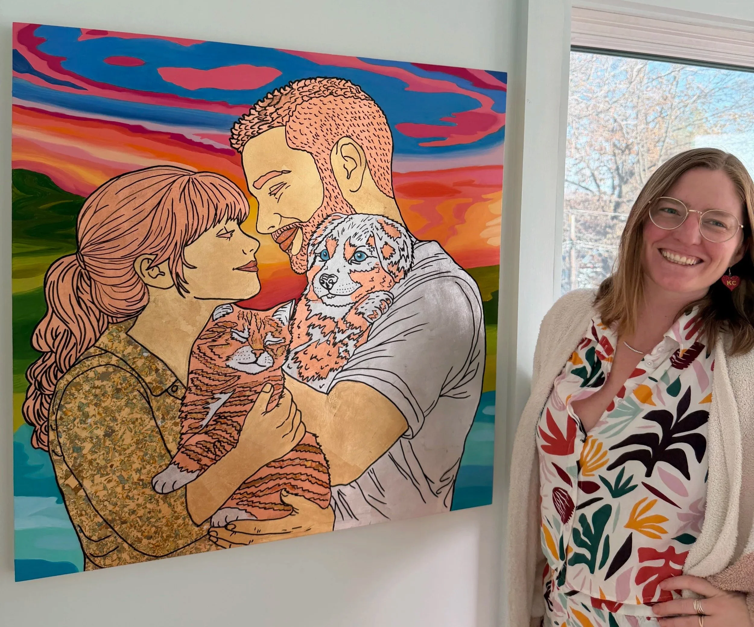 Photo of a woman standing next to an artwork featuring a man and woman holding two puppies, with a colorful sunset background.