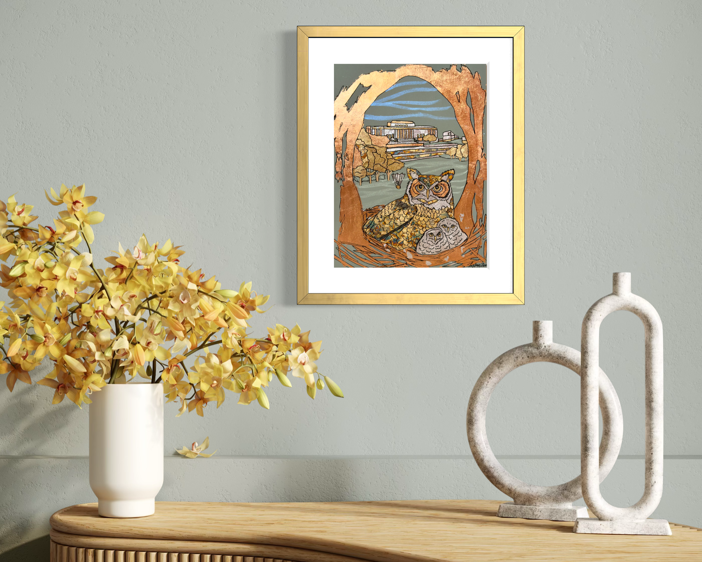 Decorative wall art depicting two owls in a nest with a cityscape and bridge in the background, placed above a wooden surface with a white vase filled with yellow and cream orchids and modern white abstract sculptures.