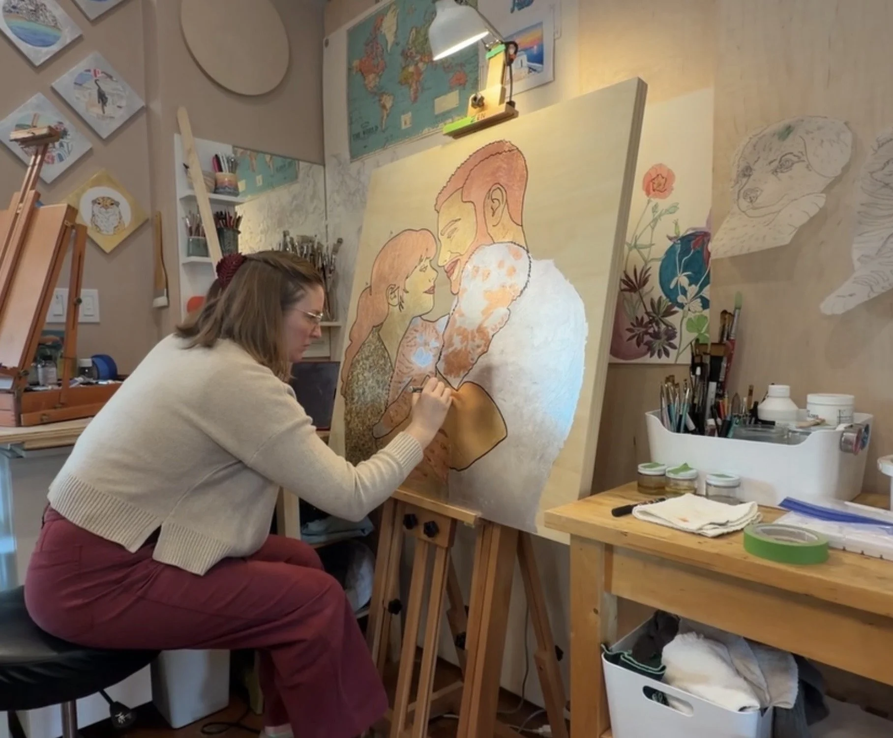 A woman sitting on a stool painting a large canvas featuring a couple embracing. The studio has art supplies, paintings on the wall, and a world map.
