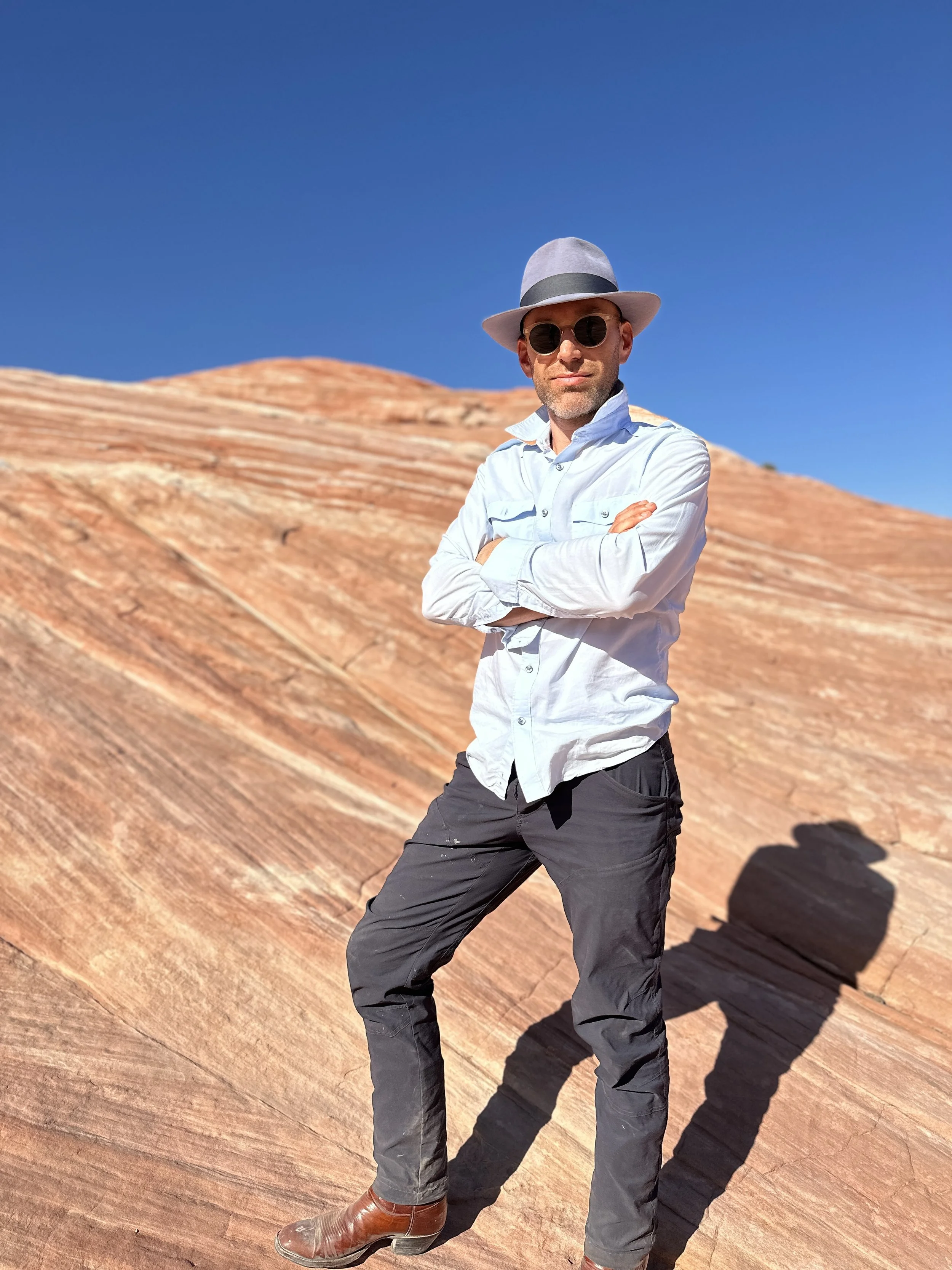 A man wearing a light gray fedora, round sunglasses, a light blue button-up shirt, black pants, and brown shoes standing with arms crossed on a rocky, reddish-brown formation under a clear blue sky.