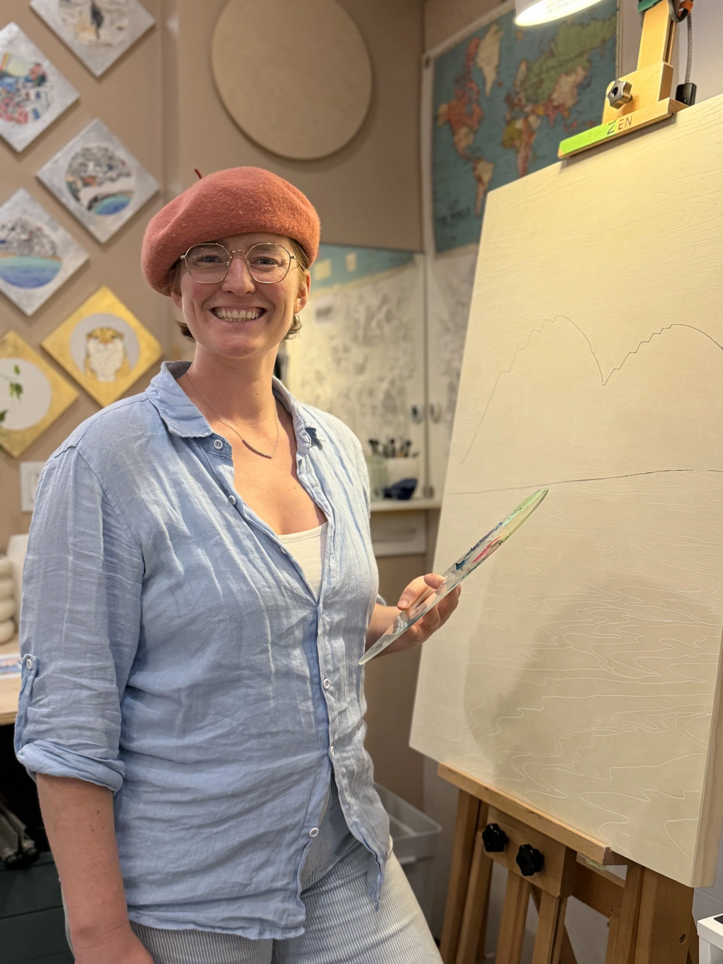 Woman smiling, wearing glasses, a pink beret, holding a palette, standing next to an easel with a landscape drawing, in an art studio with paintings on the wall and a world map in the background.