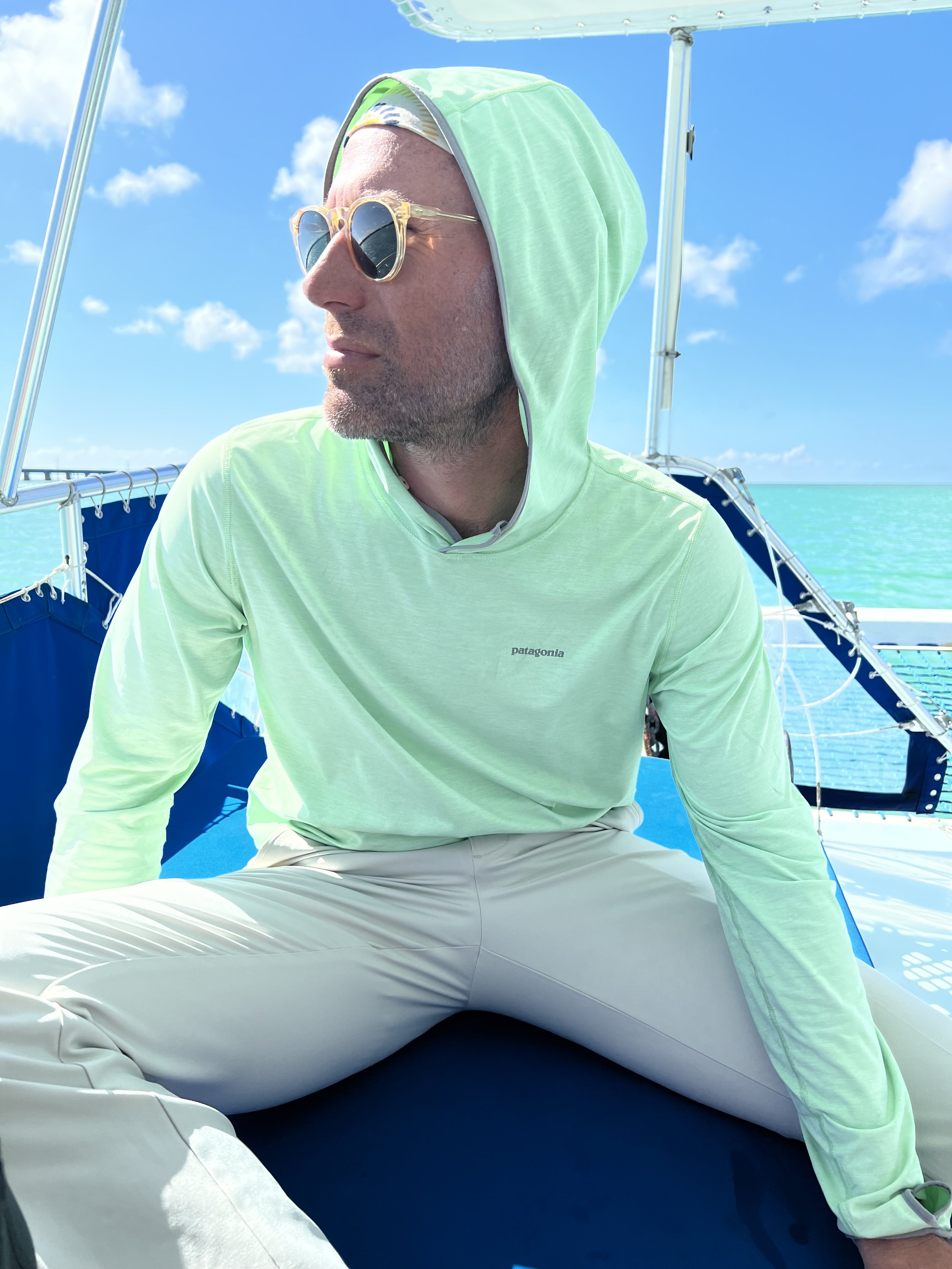 Man sitting on a boat wearing sunglasses and a light green hoodie, with the ocean and a blue sky with clouds in the background.