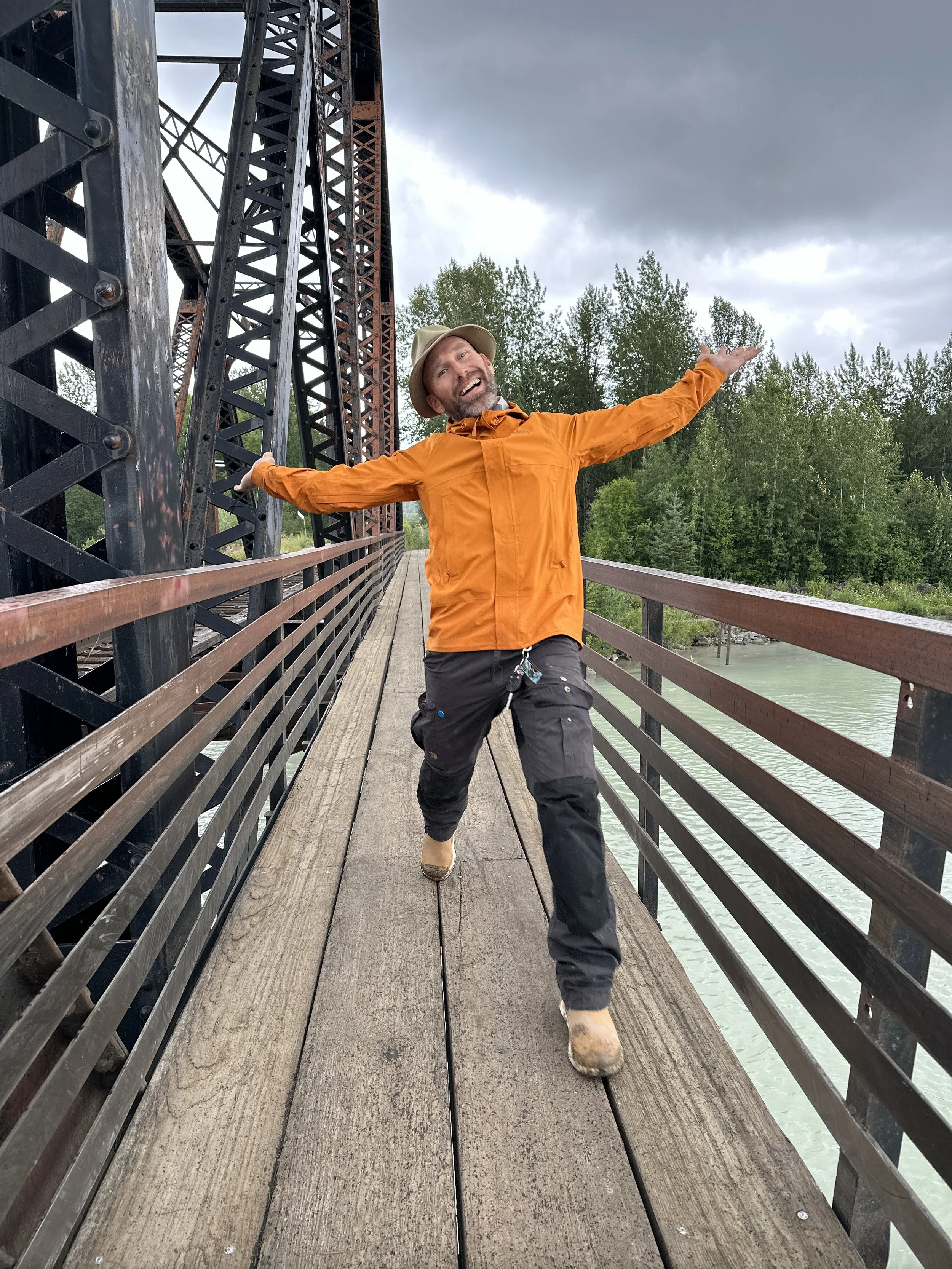 Man in an orange jacket and beige hat standing on a wooden bridge with arms outstretched and smiling, with trees and cloudy sky in the background.