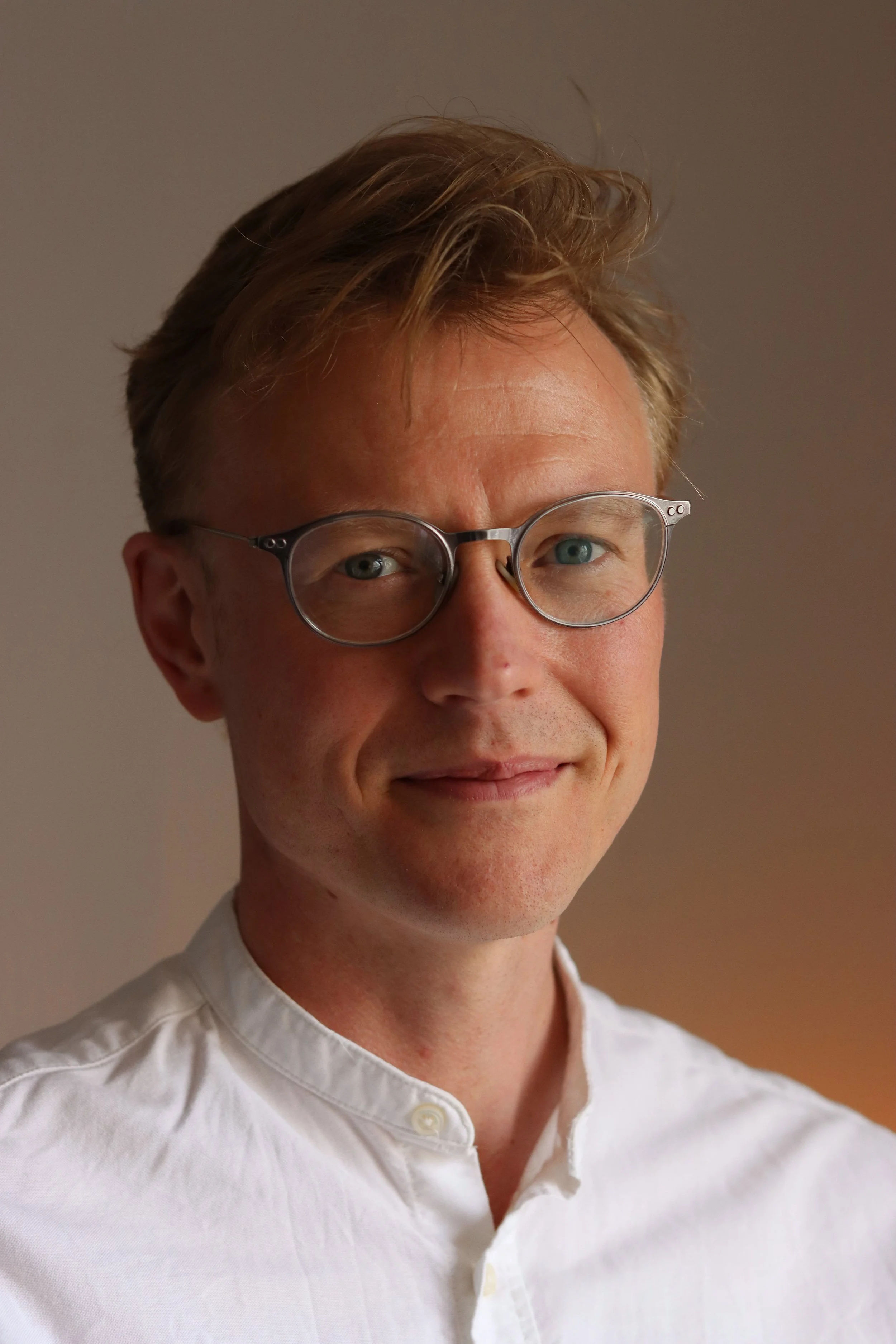 Close-up of a man with light brown hair, glasses, and a white collared shirt, smiling slightly, against a neutral background.