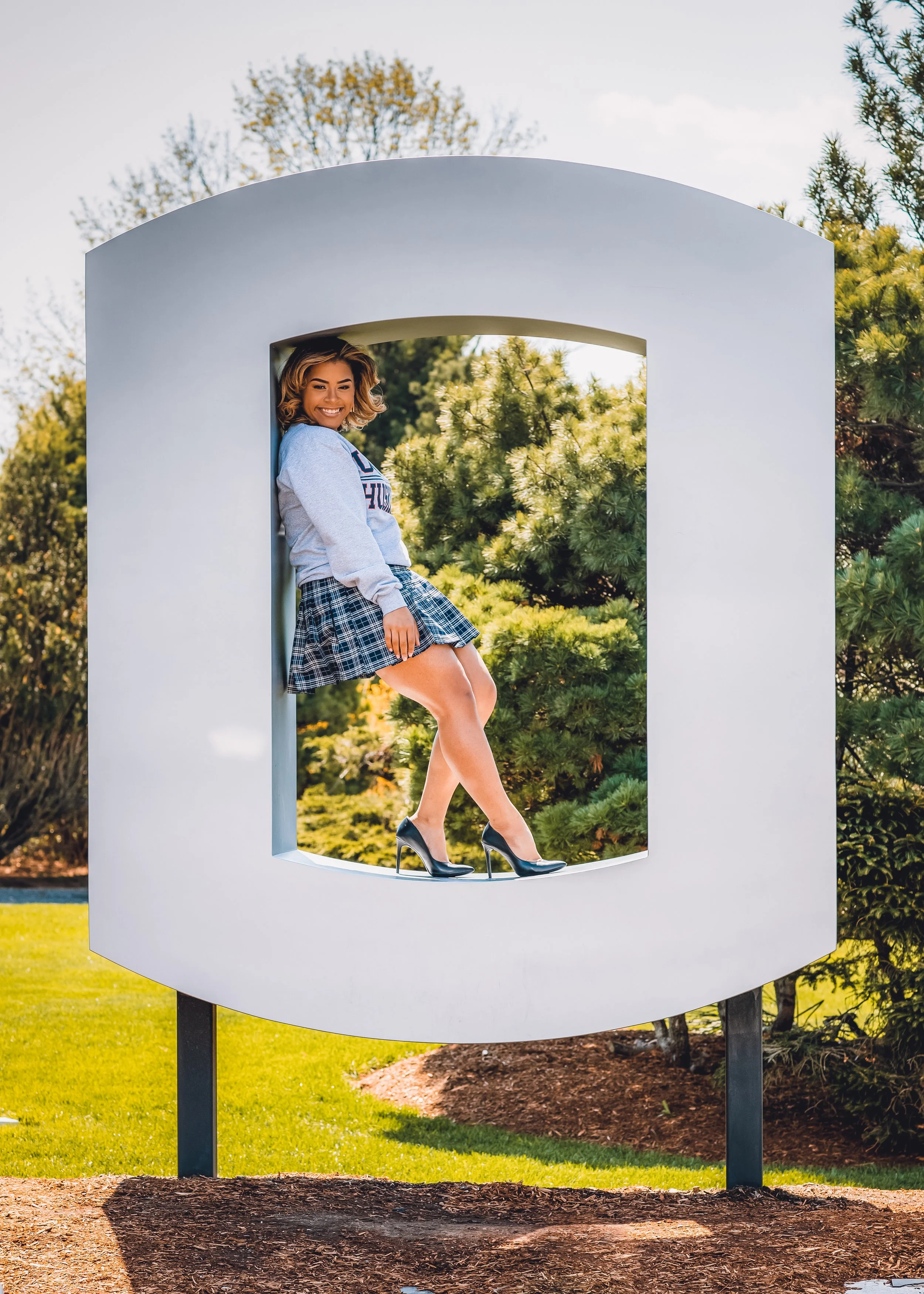 A woman with blonde hair, smiling, sitting inside a large, white, rectangular frame outside on a sunny day. She is wearing a gray sweatshirt with a logo, a plaid skirt, and black high heels. Green trees and grass are visible in the background.