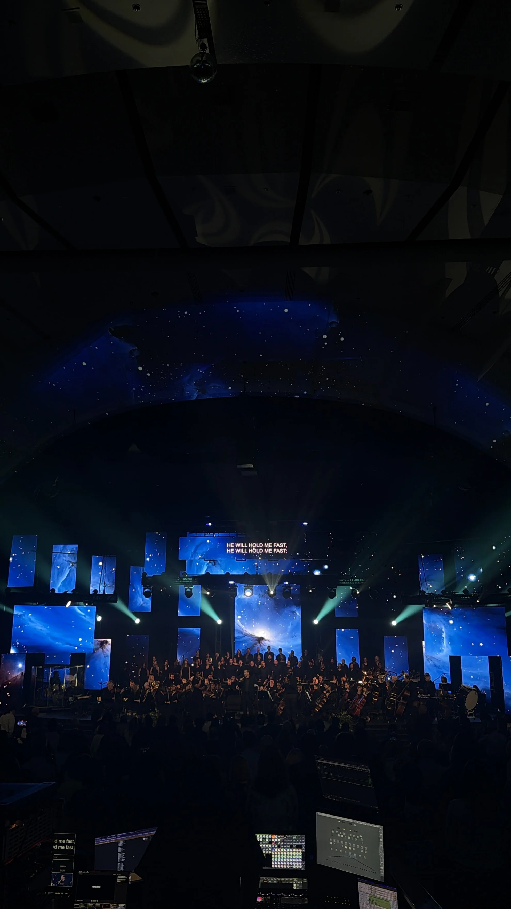 A large concert hall with a choir, orchestra, and stage lighting. The ceiling and walls are decorated with stars and space-themed visuals. A choir and orchestra are performing, with singing displayed on a large screen reading 'He will hold me fast.' Computers and sound equipment are visible in the foreground.