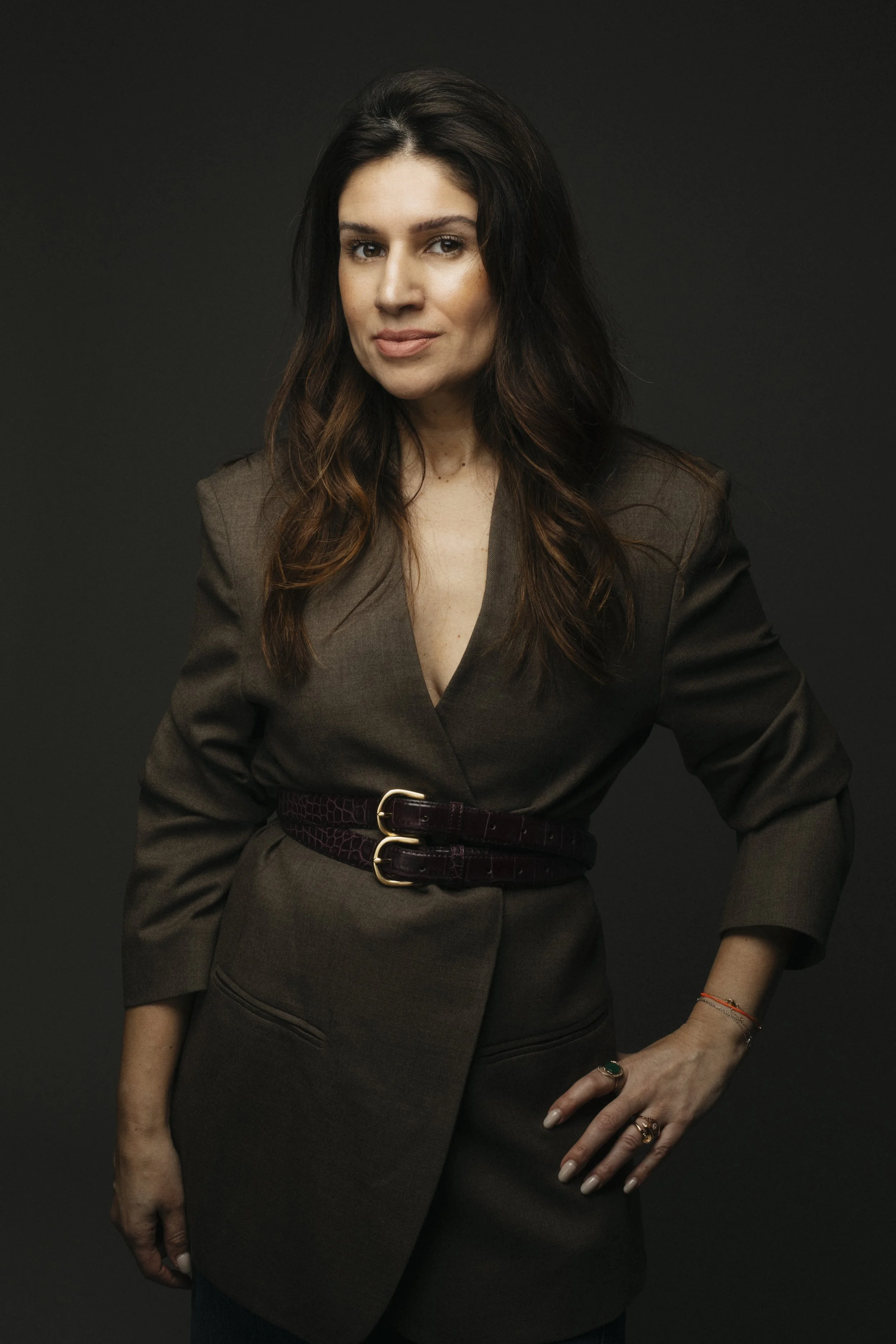 A woman with dark hair wearing a brown blazer and skirt, accessorized with rings and a bracelet, posed against a black background.