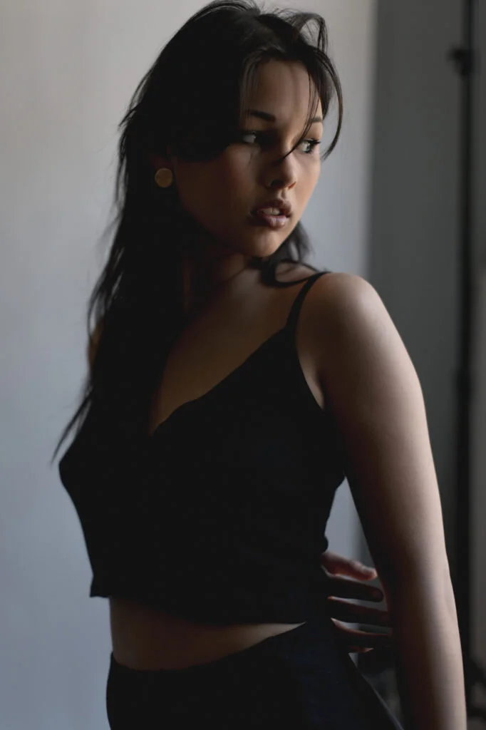 A young woman with dark hair and light skin standing near a window, wearing a black sleeveless top and looking to her right.