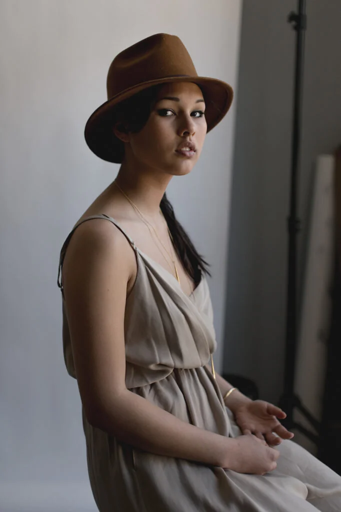 A young woman wearing a brown hat and a beige sleeveless dress, sitting indoors against a plain white wall.