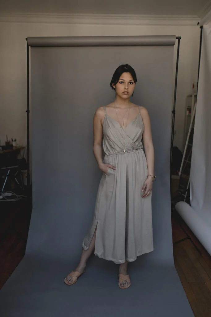 Young woman in a light gray dress and sandals standing in front of a gray photo backdrop in a studio.