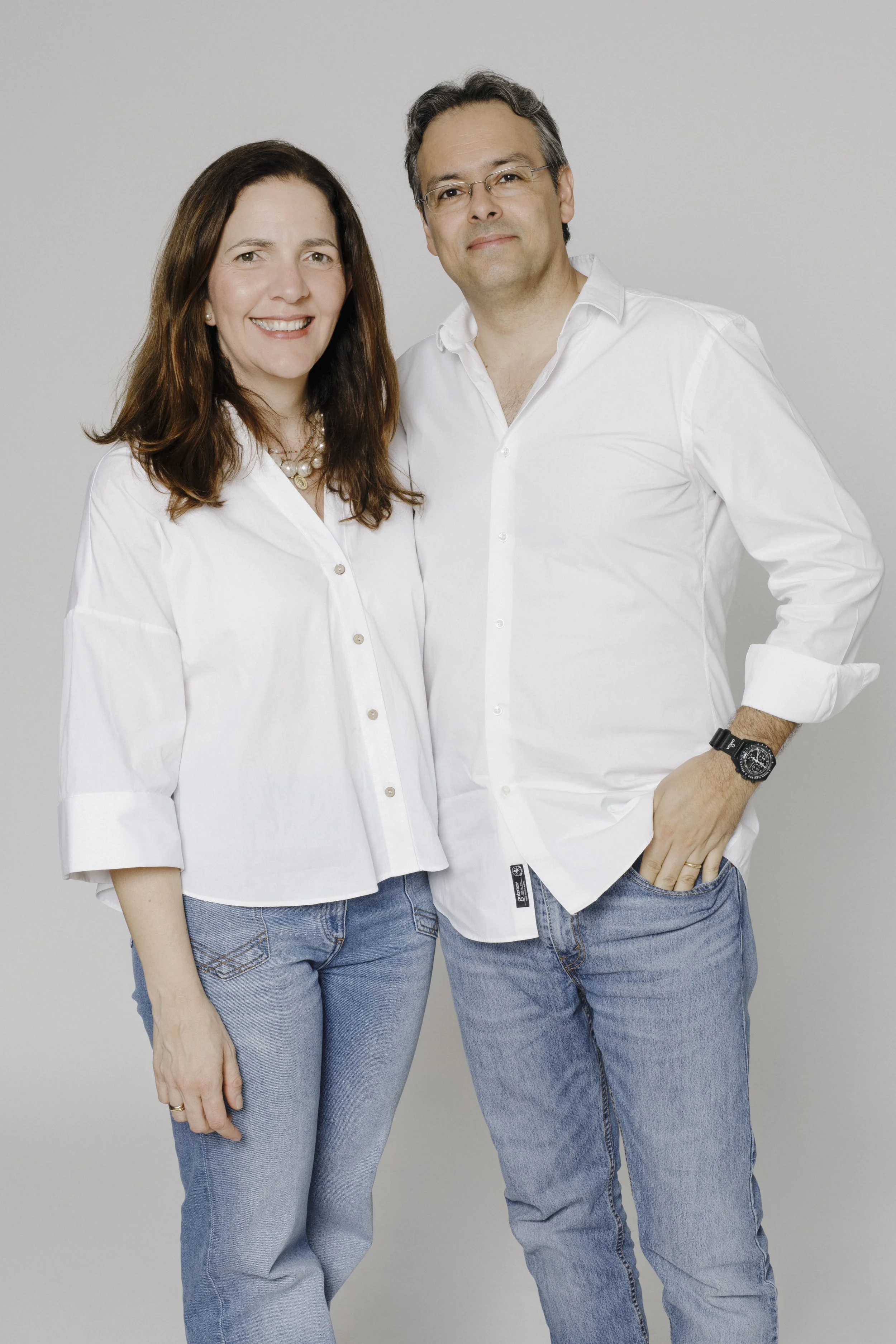 A woman with long brown hair wearing a white button-up shirt and jeans, standing next to a man with short gray hair and glasses, wearing a white button-up shirt and jeans, against a plain light gray background.