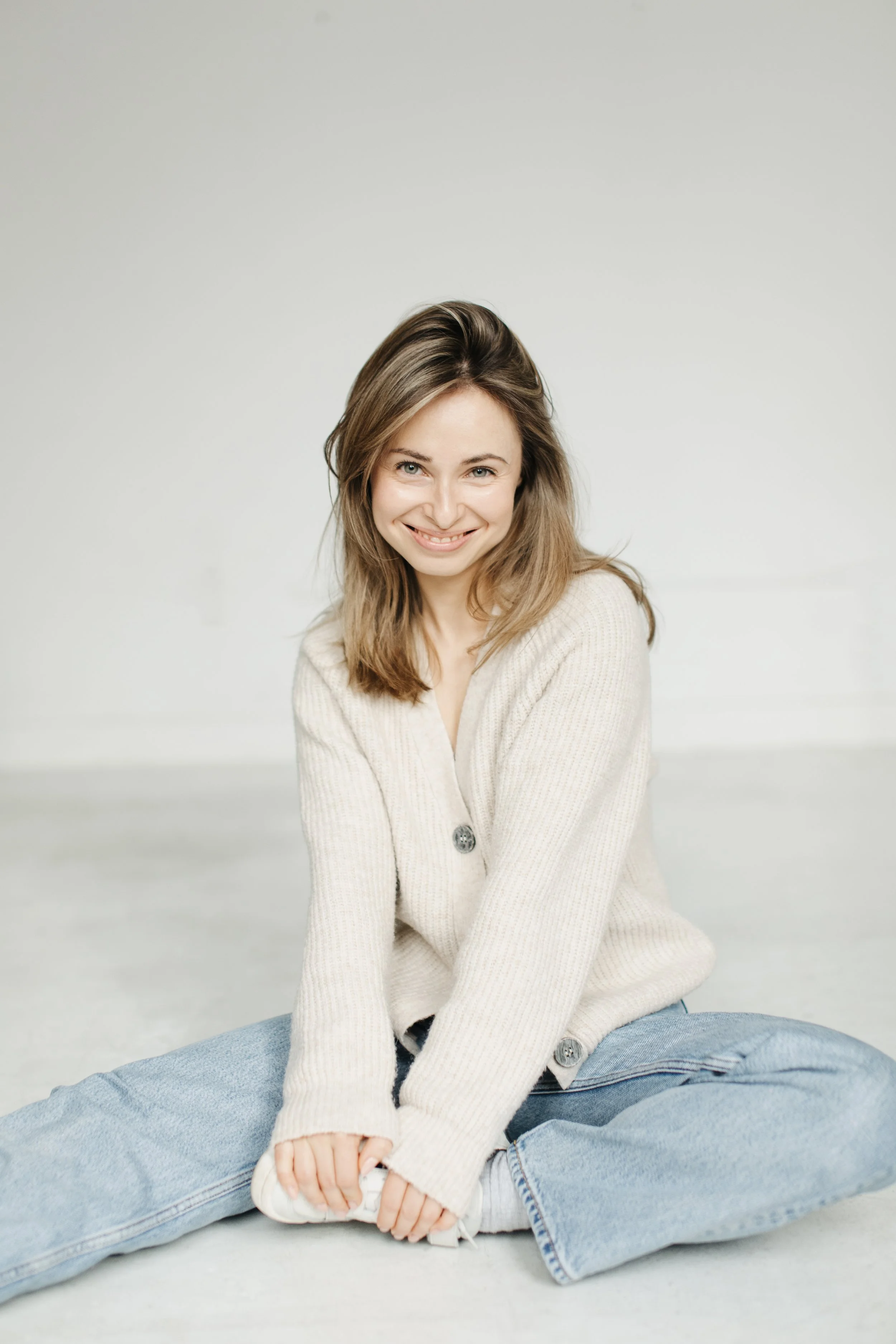 A young woman with long brown hair sitting on the floor, smiling, wearing a cream-colored cardigan and light blue jeans, against a plain light background.