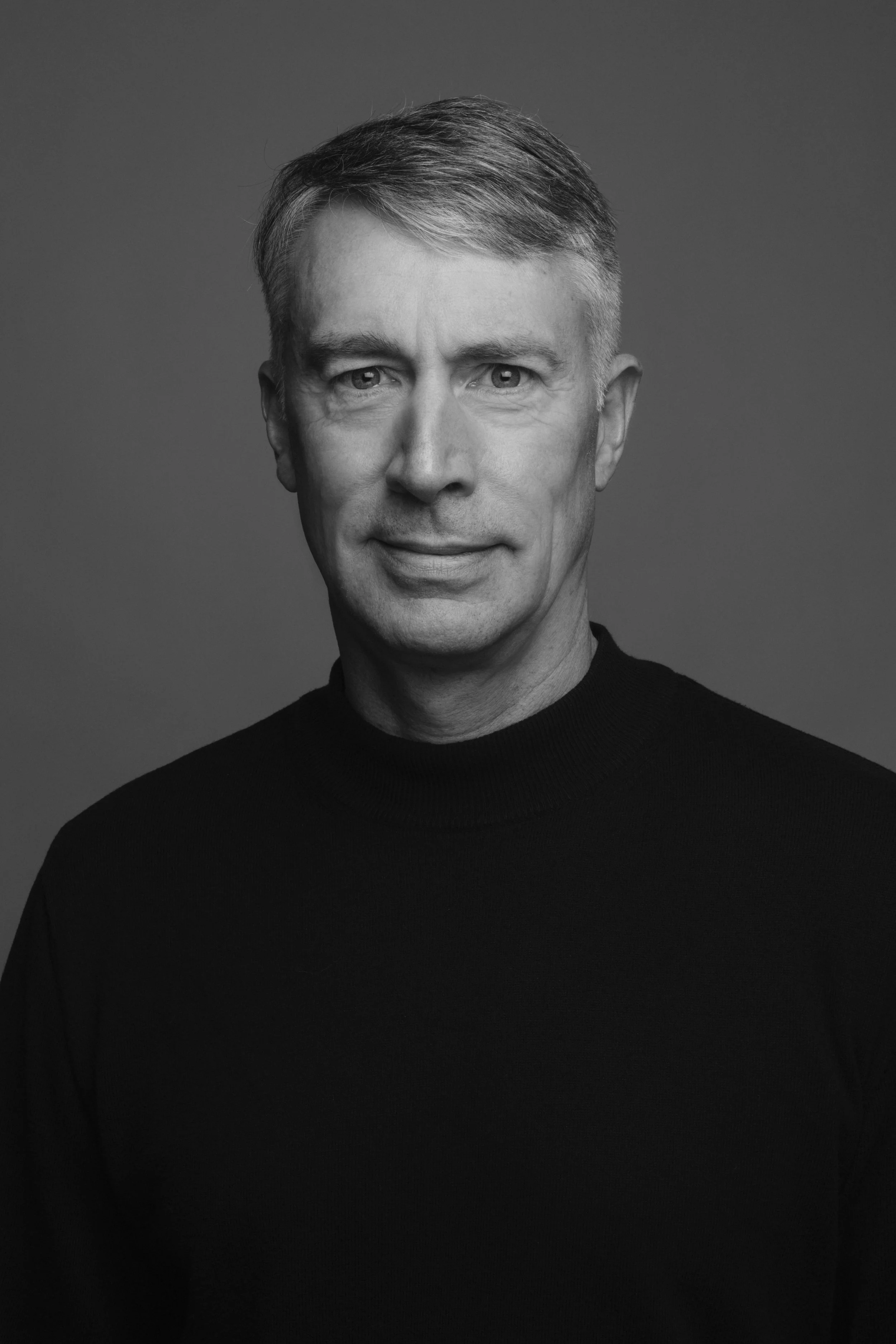 Black and white portrait of a middle-aged man with short, neatly styled hair, wearing a black turtleneck sweater, looking directly at the camera with a slight smile, against a plain background.