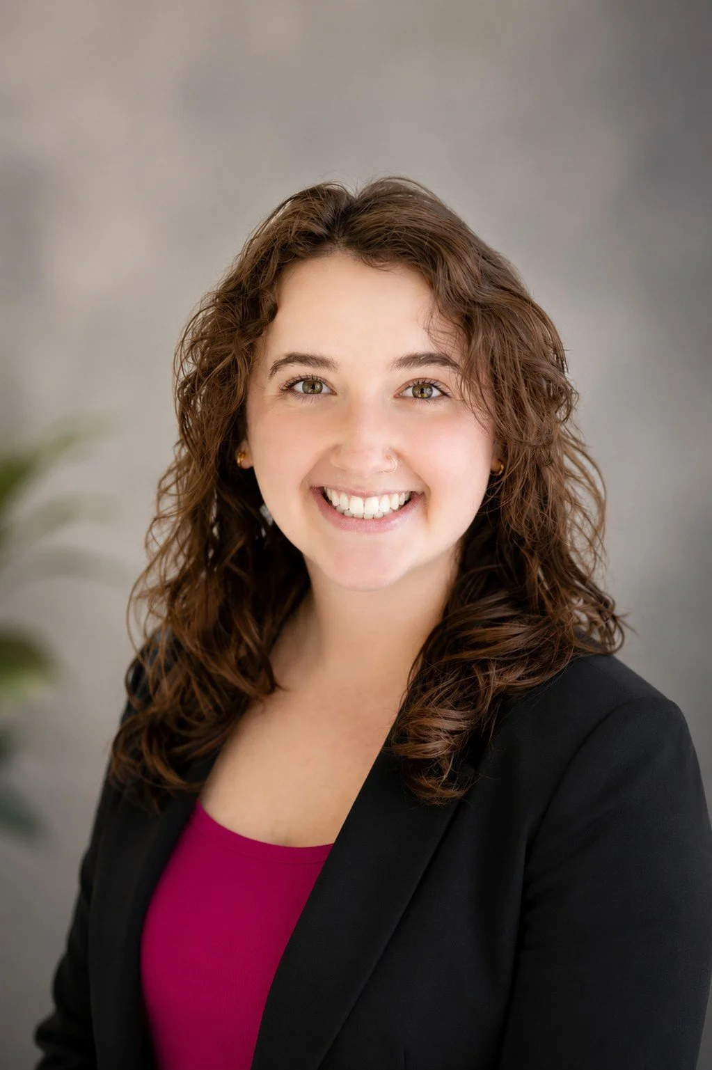 A woman with curly brown hair, hazel eyes, and a nose piercing, smiling at the camera, wearing a magenta top against a white background.