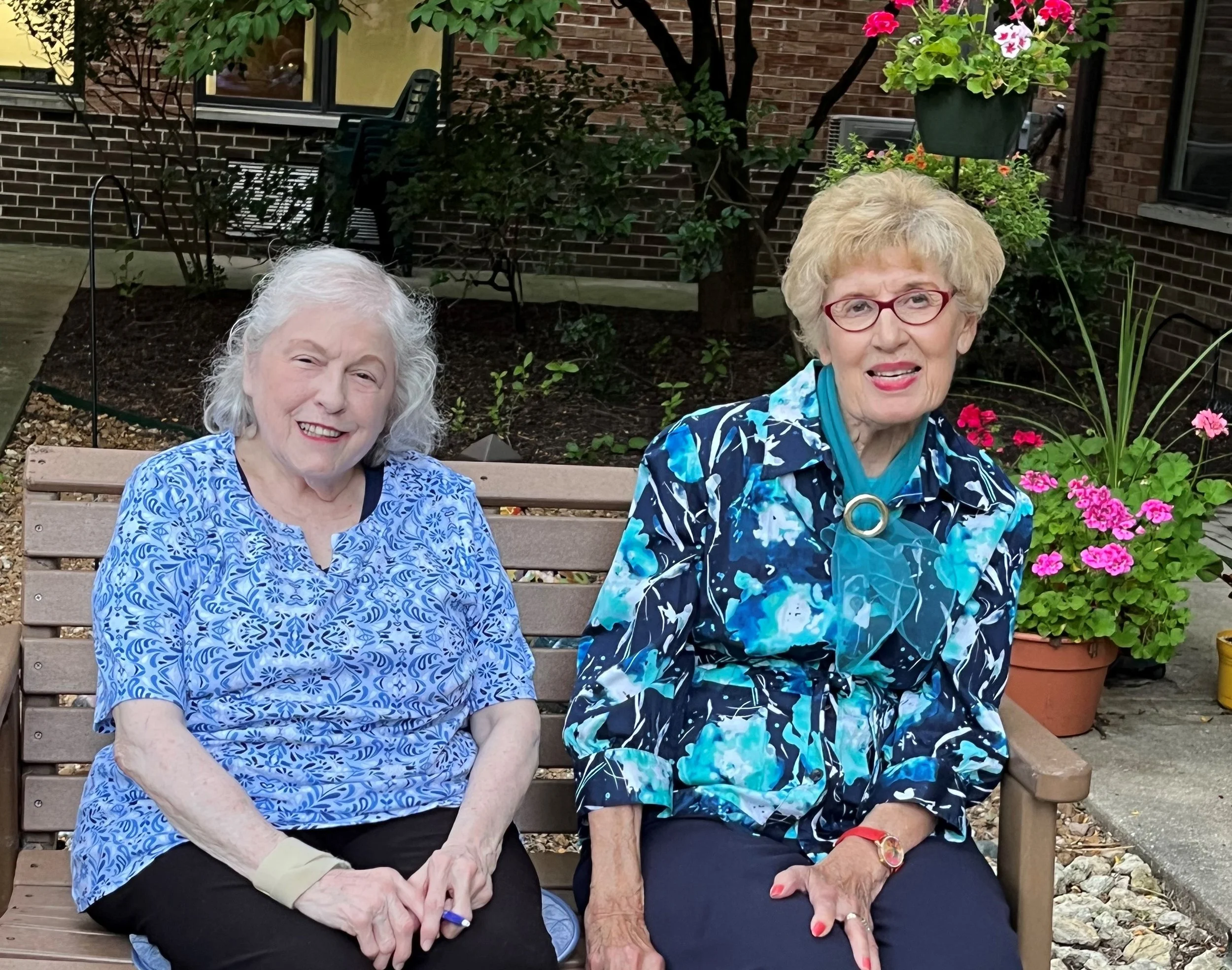 Two senior women smiling while sitting on an outdoor bench at Siena on Brendenwood, surrounded by trees and landscaping, representing the welcoming community and friendships available at our independent senior living community in Rockford, Illinois.