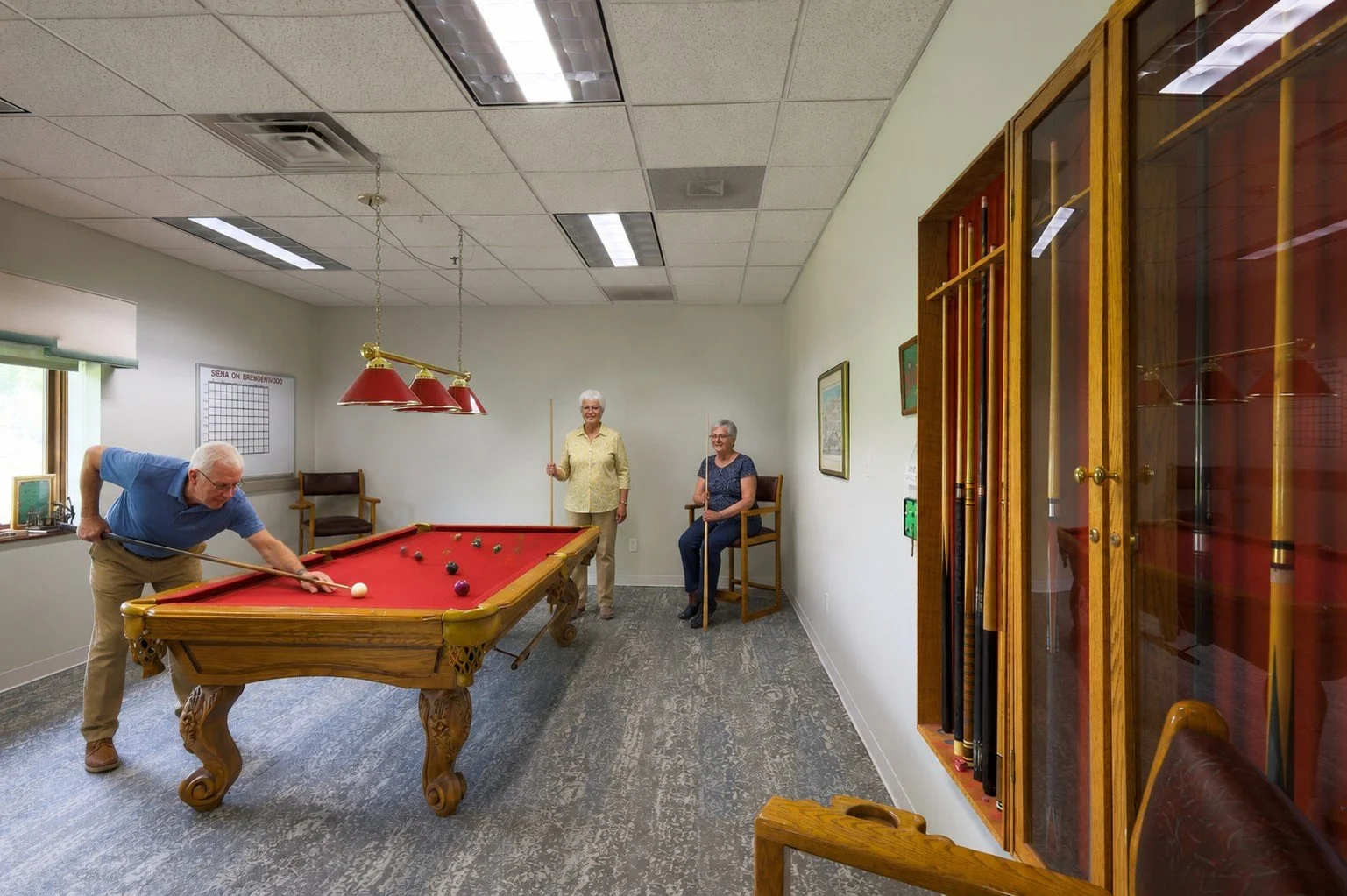 Three elderly women and one elderly man playing billiards in a room with pool cues and artwork on the wall.