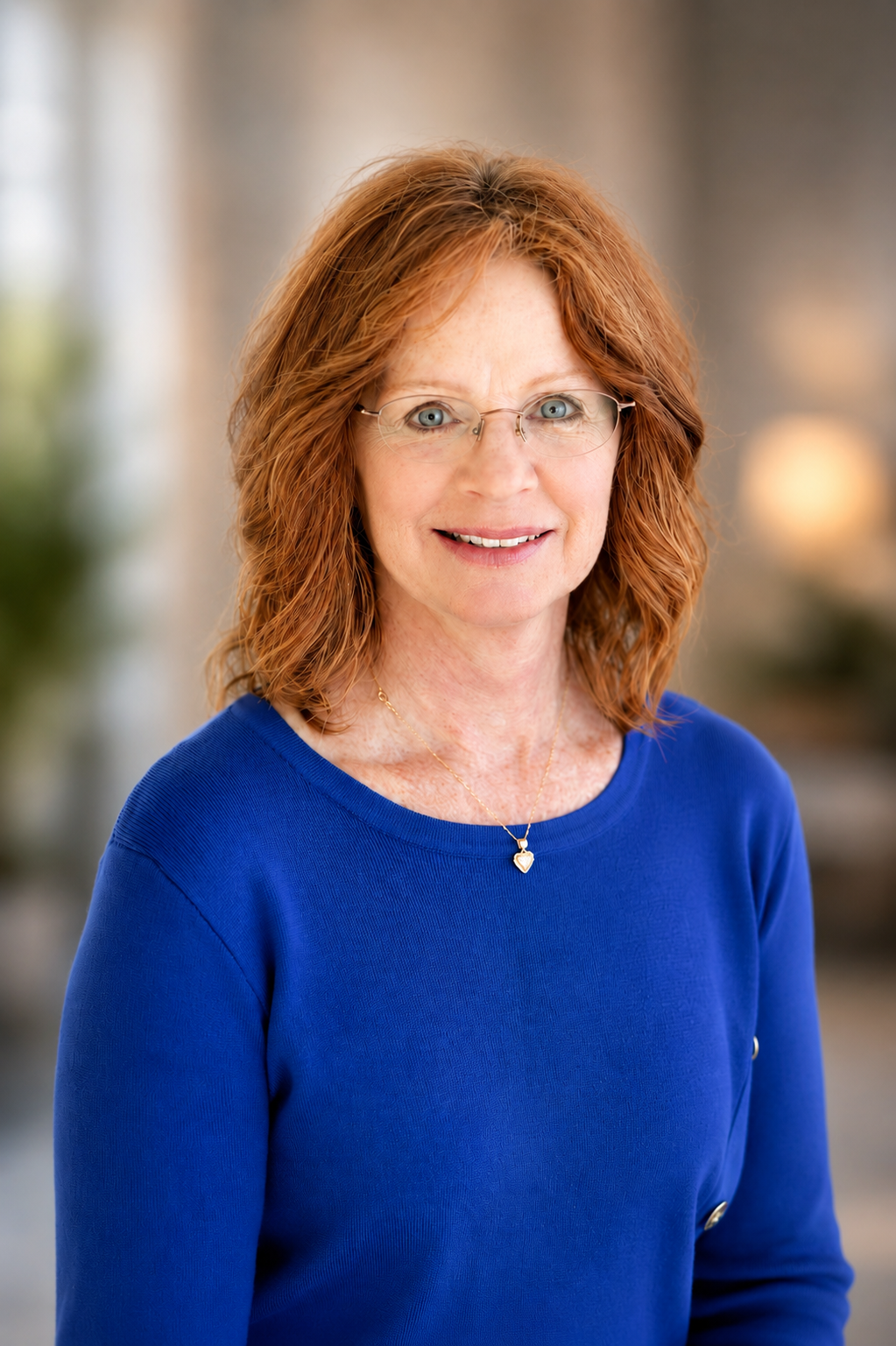 A smiling middle-aged woman with red hair, glasses, and a necklace, wearing a bright blue top, against a plain white background.
