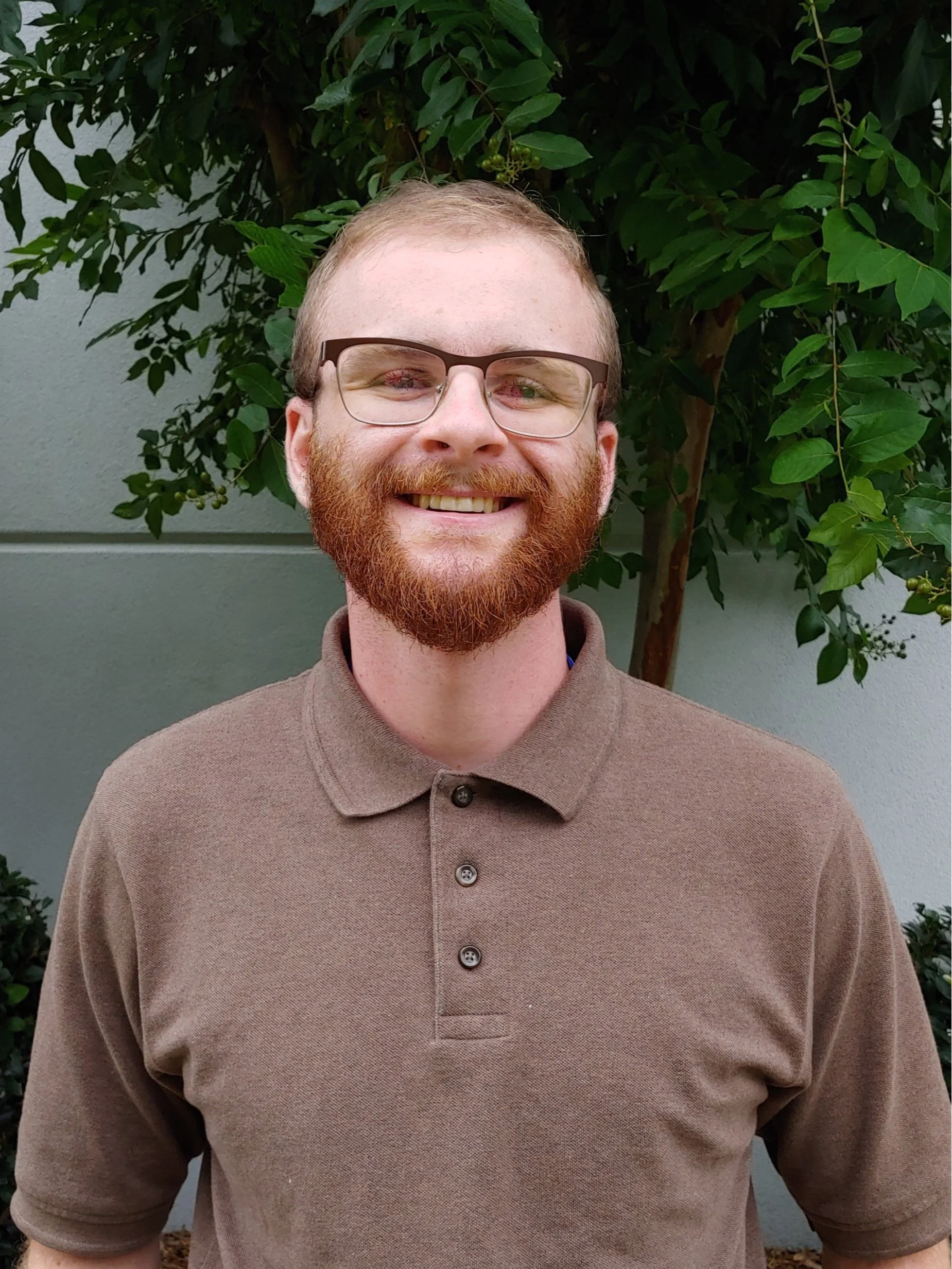 A smiling man with glasses and a red beard is standing outdoors in front of green foliage and a light gray wall.