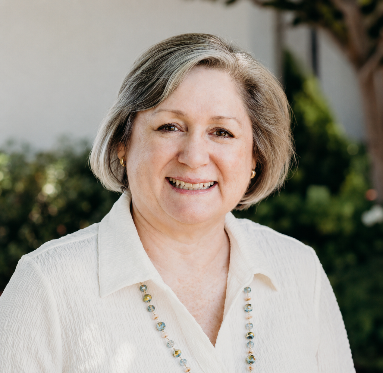 A smiling middle-aged woman with short, gray hair, wearing a white blouse, gold earrings, and a beaded necklace, standing outdoors with greenery in the background.