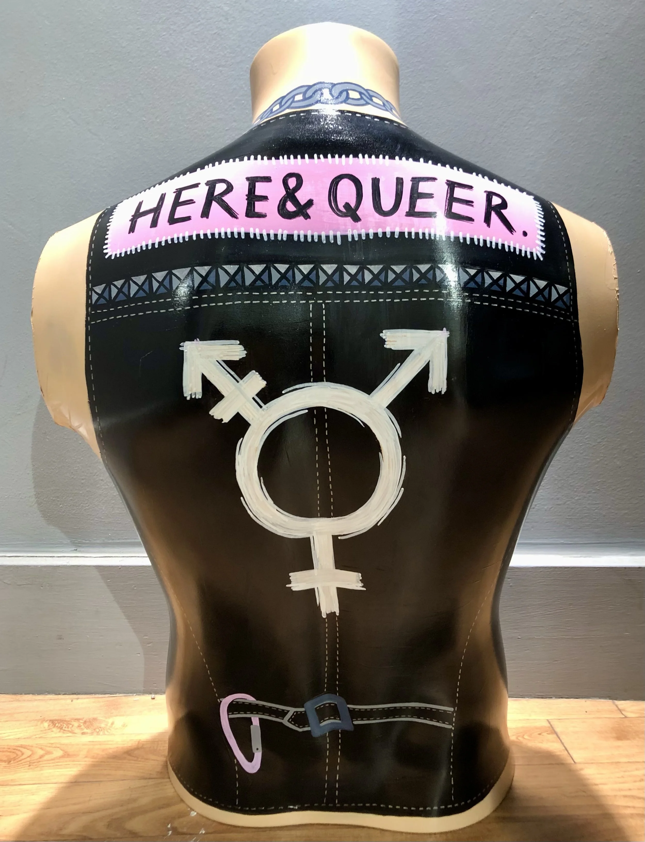 The back of a male mannequin torso which has been painted to look like it is wearing a leather vest. The vest includes a patch that says Here & Queer, studs along the seam, a trans pride icon, and a lesbian carabiner. The artwork is by Hayley Wells