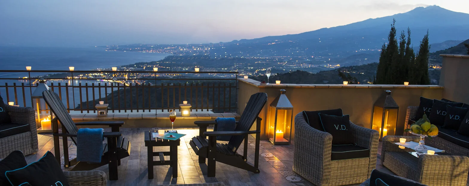 A rooftop terrace at dusk with outdoor furniture, lanterns, candles, and a scenic view of a coastal city and mountains in the background.