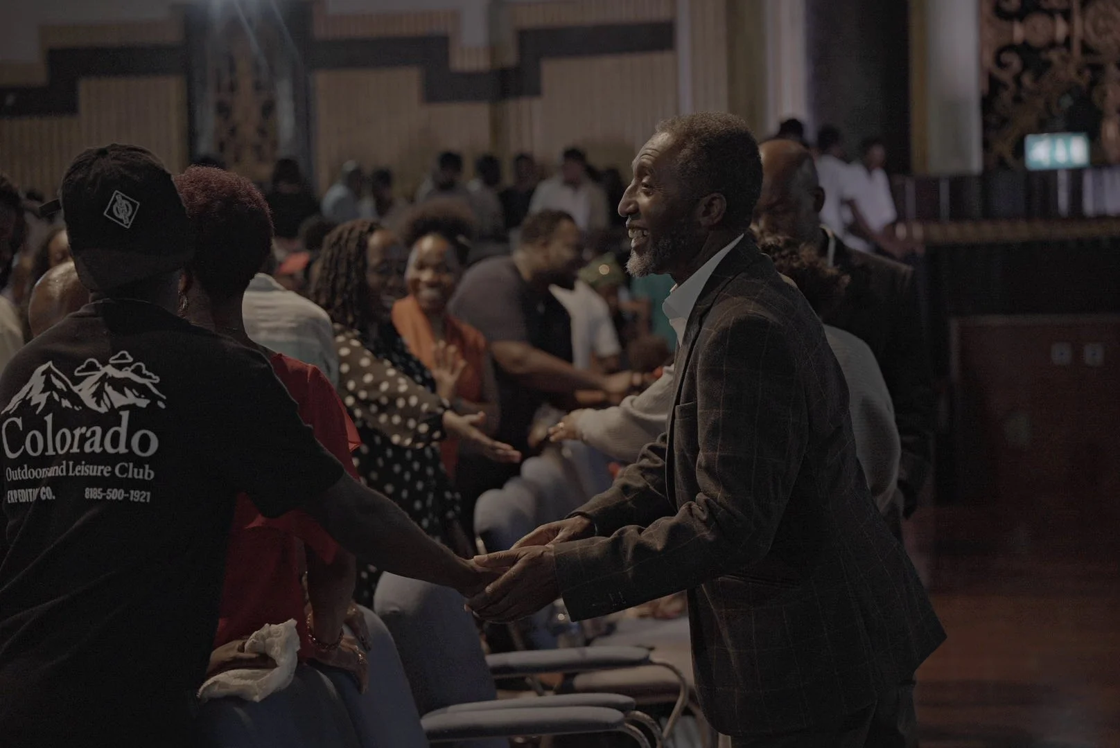 A man and woman holding hands and smiling during a gathering in a dimly lit room, with other people seated and standing in the background.
