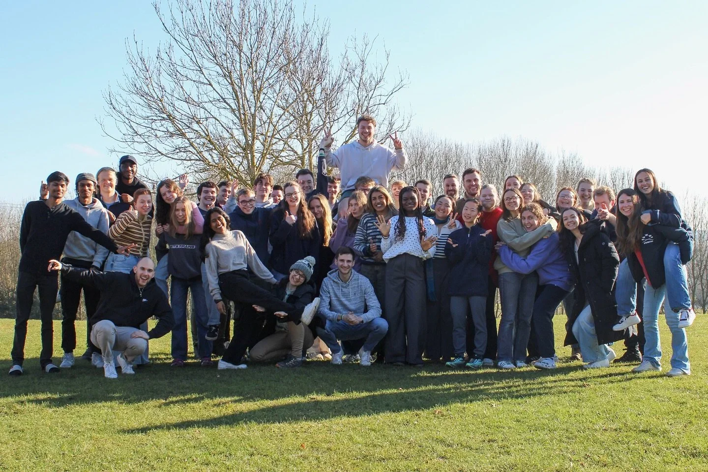 A large group of diverse young people smiling and posing outdoors on a sunny day in a grassy field with leafless trees in the background.