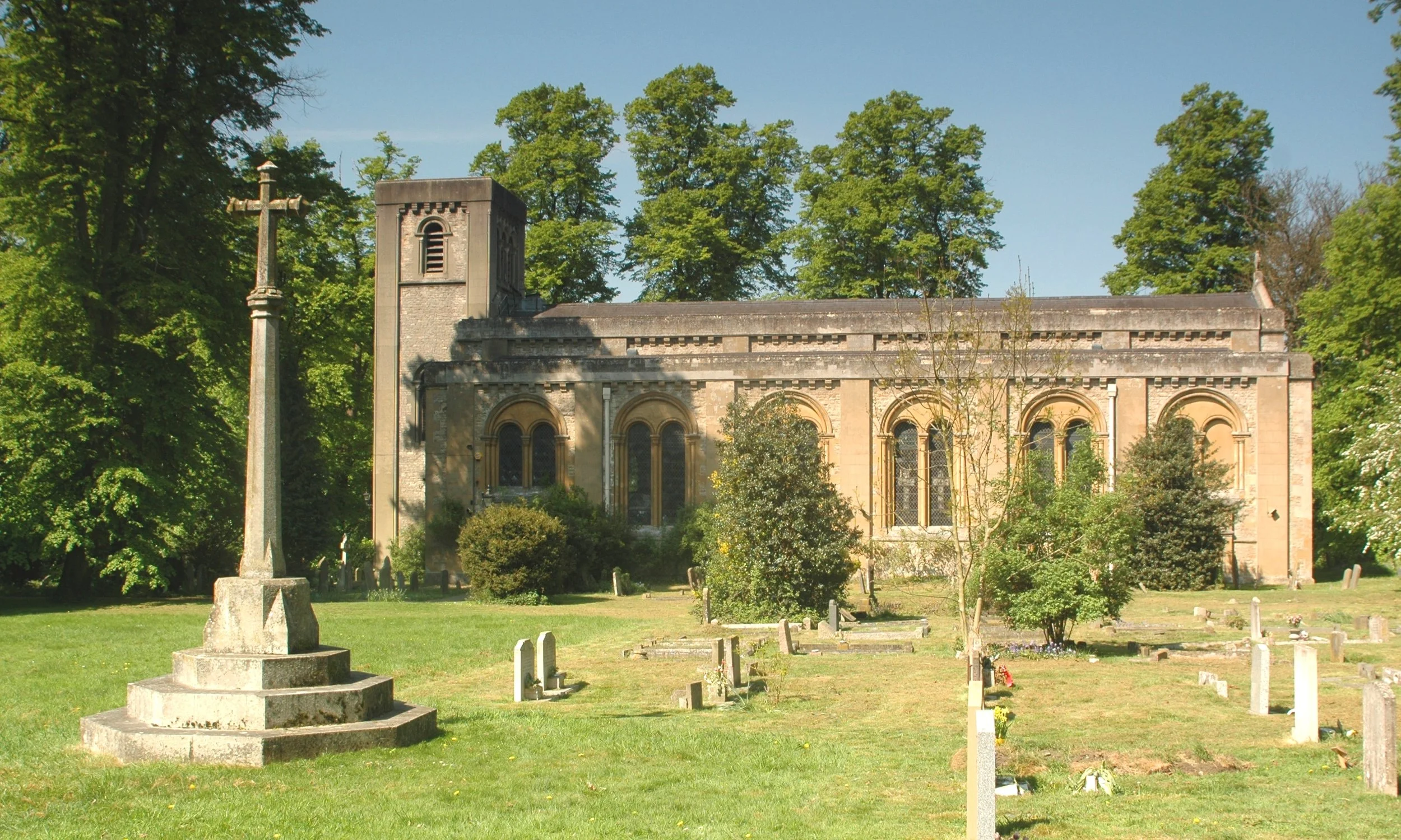 A stone church with arched windows surrounded by trees and a graveyard, with a tall stone cross monument in the foreground.