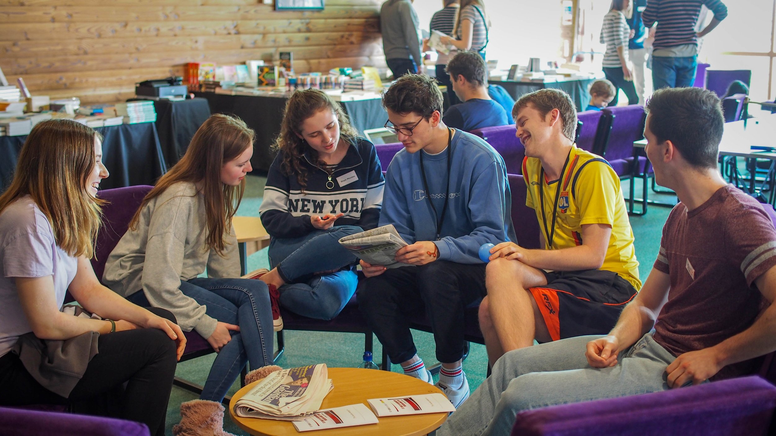Group of young people sitting in a circle, engaged in conversation and smiling, at an indoor event with tables and books in the background.