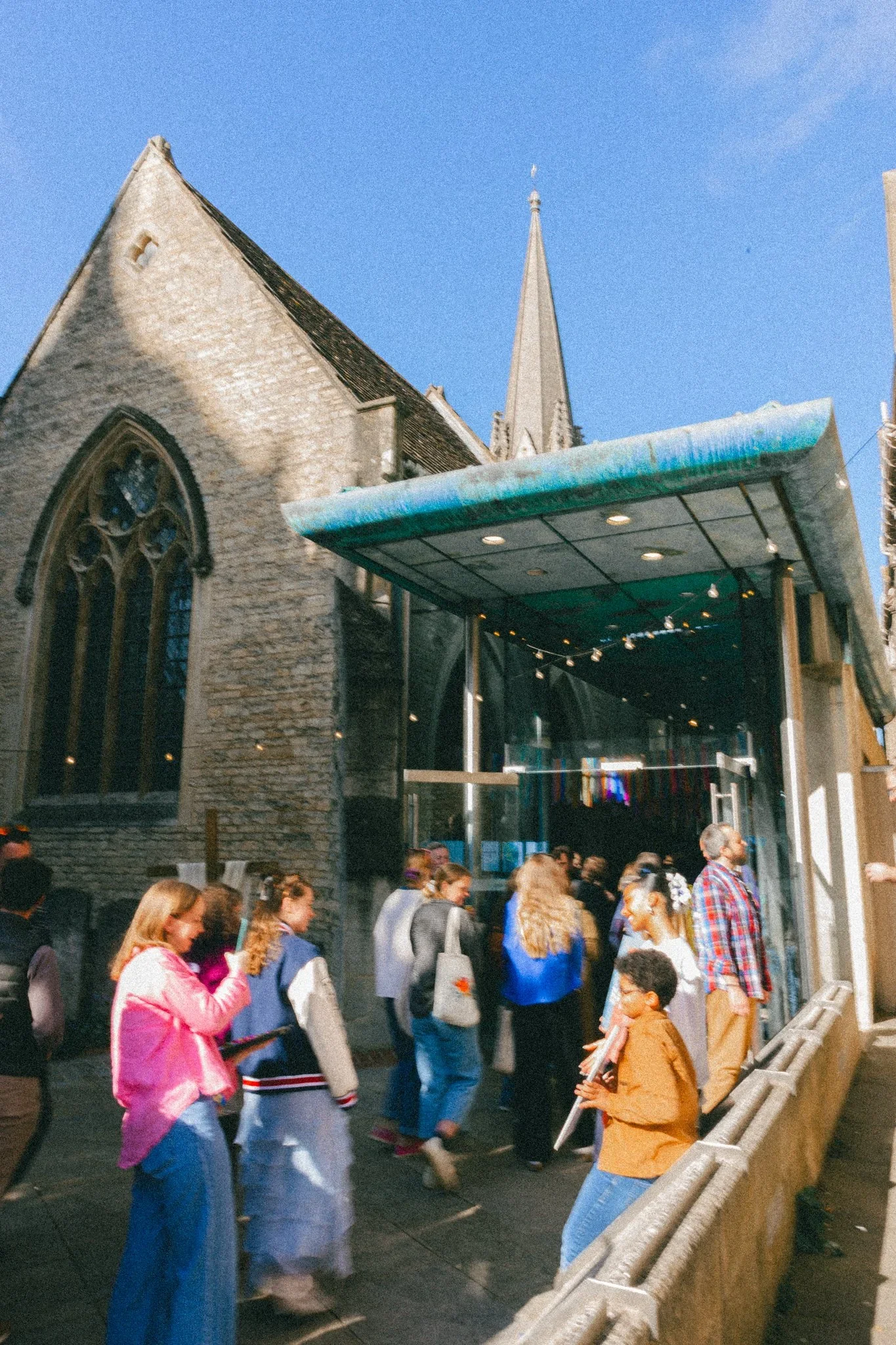 People queuing outside a church with a modern glass entrance in an urban setting on a sunny day.